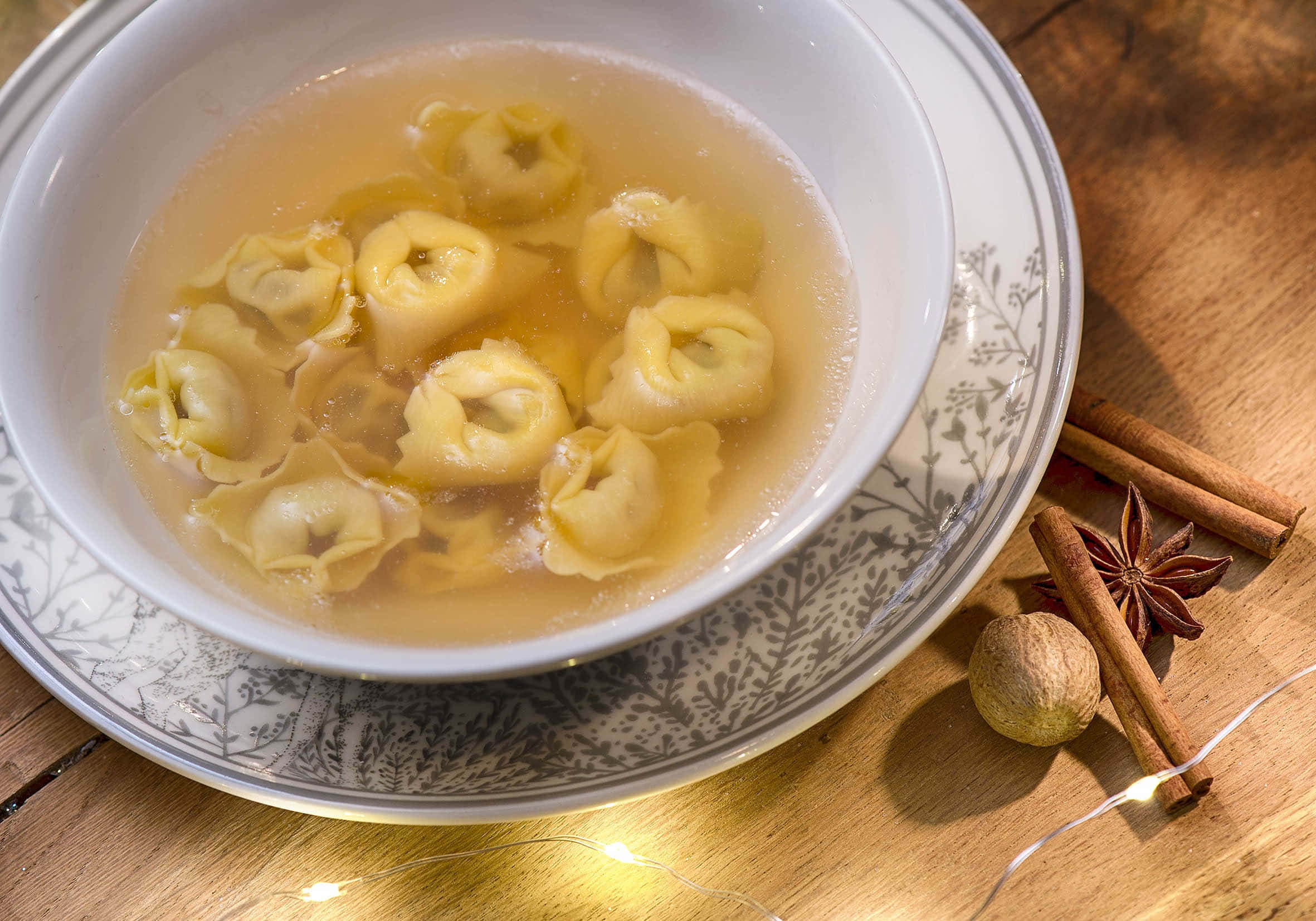 Food Photography 桌布 壁紙 — 一碗熱騰騰的義大利餃湯放在有圖案的盤子上，旁邊有肉桂棒、八角茴香和肉豆蔻，放在一個有仙女燈的木製桌子上。