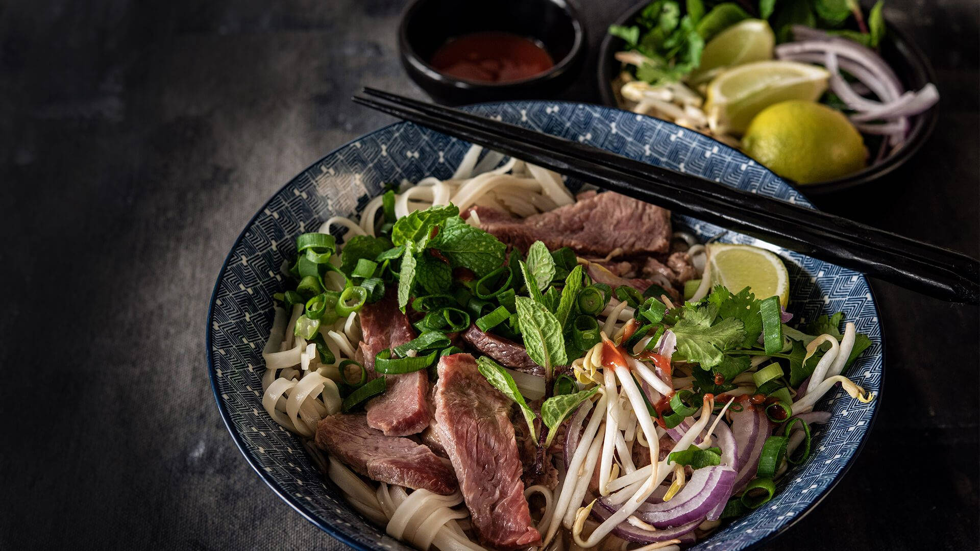 Food Photography 桌布 壁紙 — 一碗熱騰騰的越南河粉，配有牛肉麵、新鮮香草、萊姆和豆芽，並以筷子點綴。