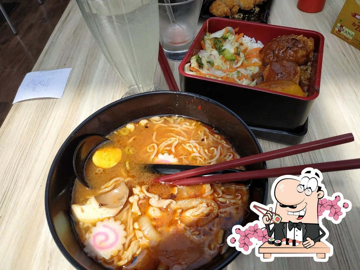 Noodles 桌布 壁紙 — 一碗熱騰騰的辛辣拉麵，配上溏心蛋和配料，旁邊放著一個便當盒，放在一張帶有卡通貼紙的木桌上。