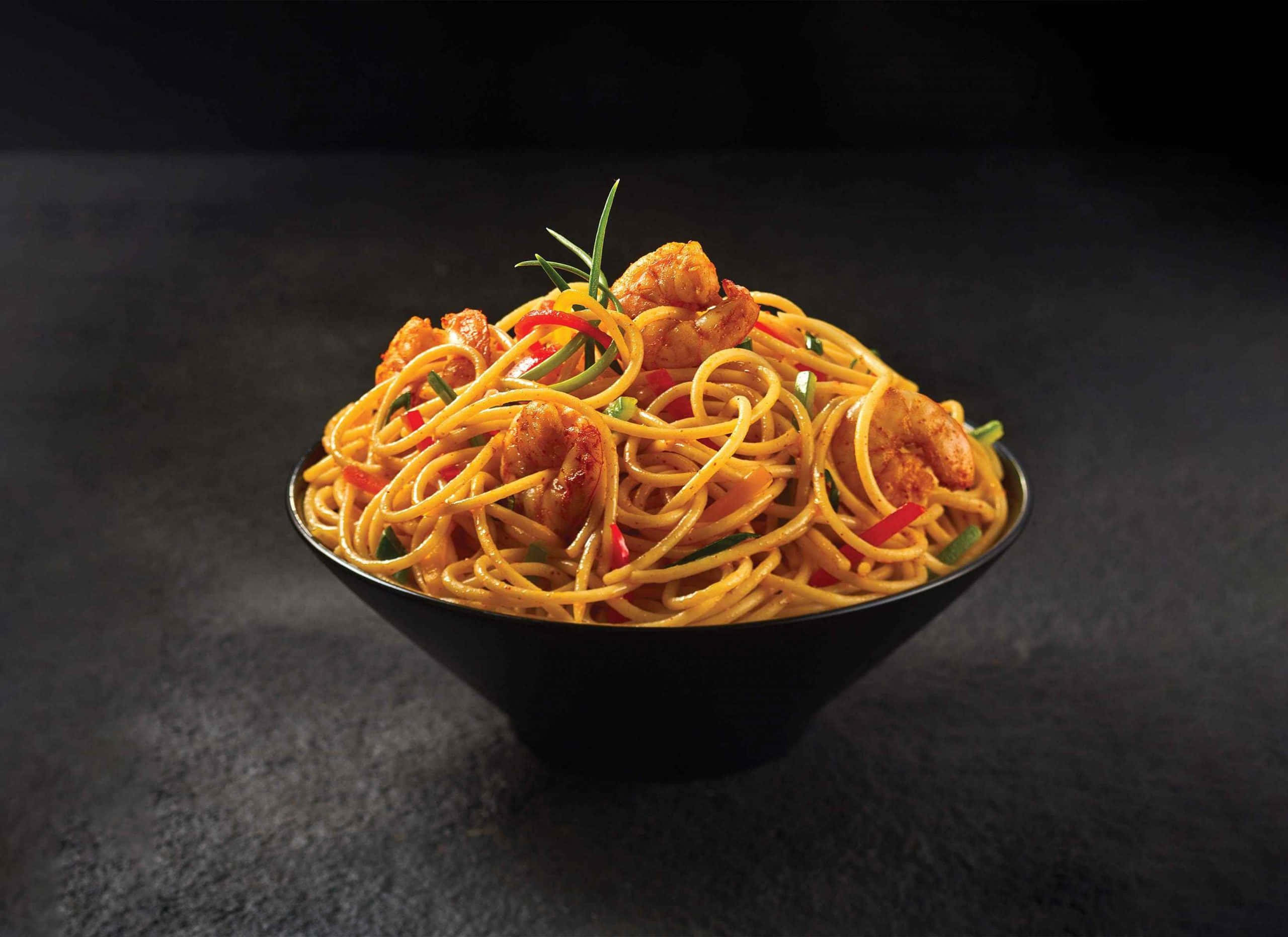 Food Photography 桌布 壁紙 — 一碗熱騰騰的金色麵條，上面鋪滿多汁的蝦和鮮豔的紅椒，襯托著深色紋理背景。