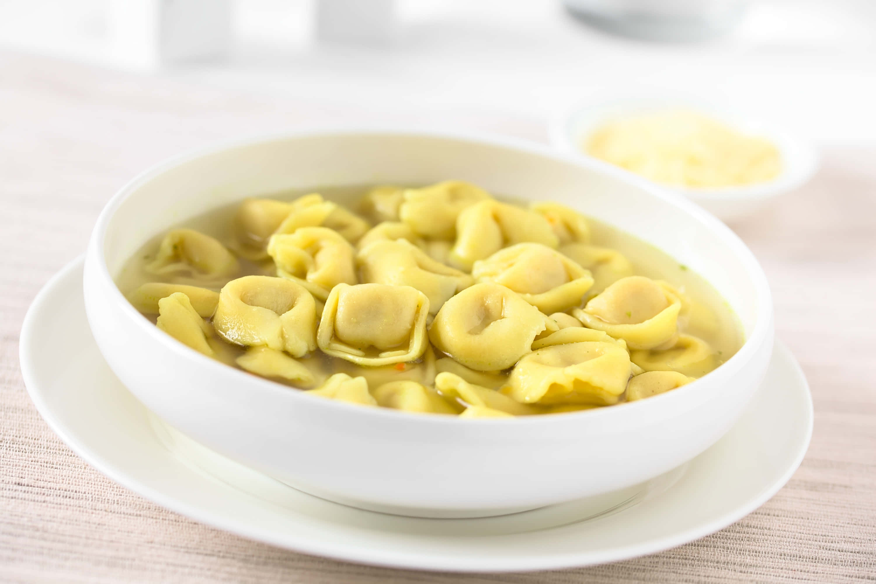 Food Photography 桌布 壁紙 — 一碗熱騰騰的金黃色義大利餃子放在清湯中，置於白色盤子上，旁邊有磨碎的起司。