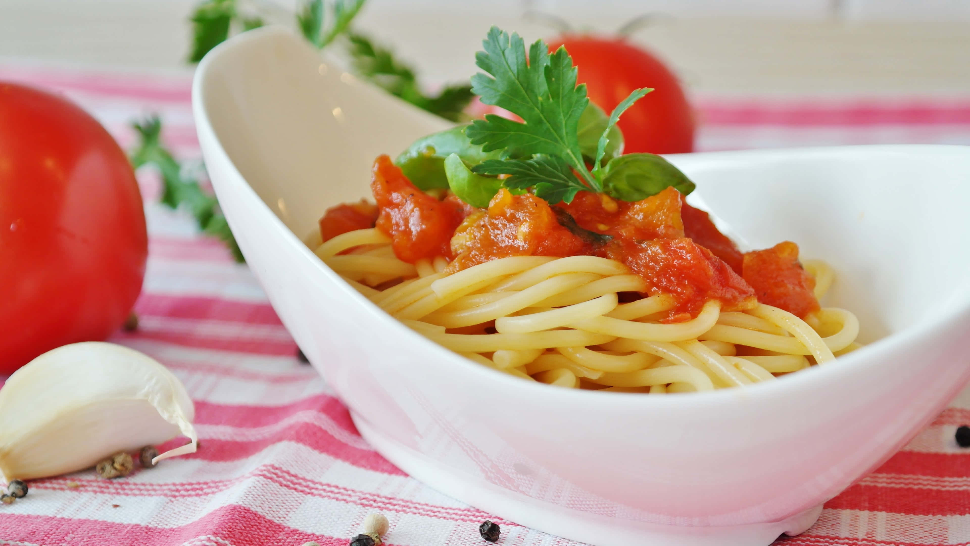 4K Pasta 桌布 壁紙 — 一碗白色義大利麵配番茄醬和新鮮歐芹，放在紅白格子布上，旁邊有整個番茄和大蒜。
