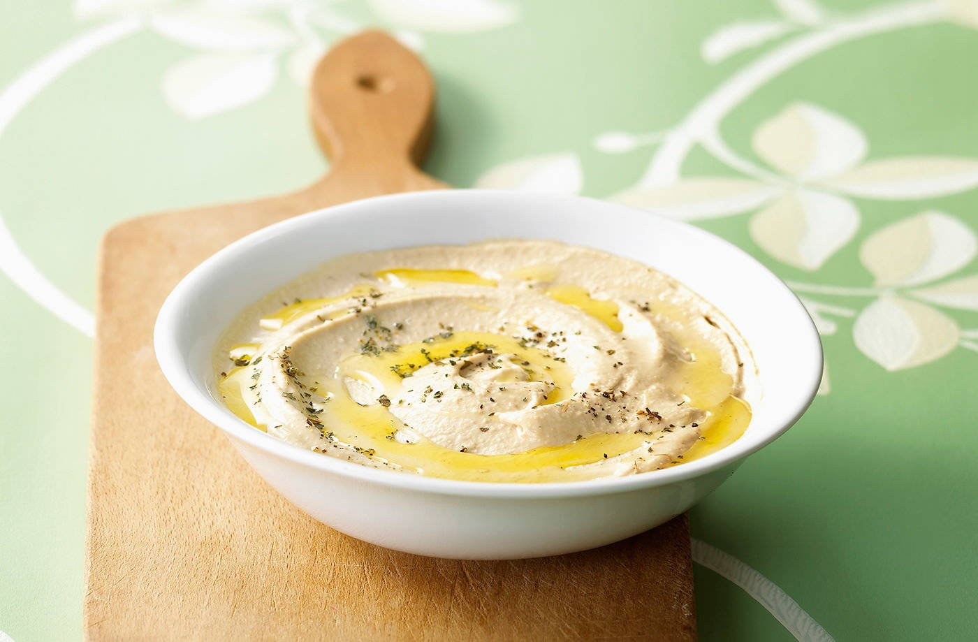 Food Photography 桌布 壁紙 — 一碗綿密的鷹嘴豆泥，淋上金黃色的橄欖油，撒上香草，放在木板上，背景是柔和的綠色花卉圖案。