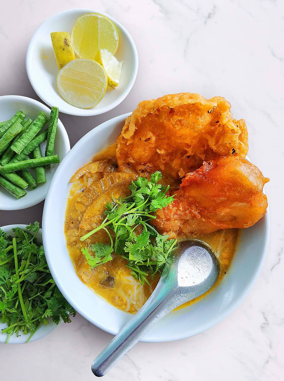 Food Photography 桌布 壁紙 — 一碗色彩繽紛的咖哩，配上炸魚和新鮮香菜裝飾，伴以檸檬角和青豆，置於大理石表面上。