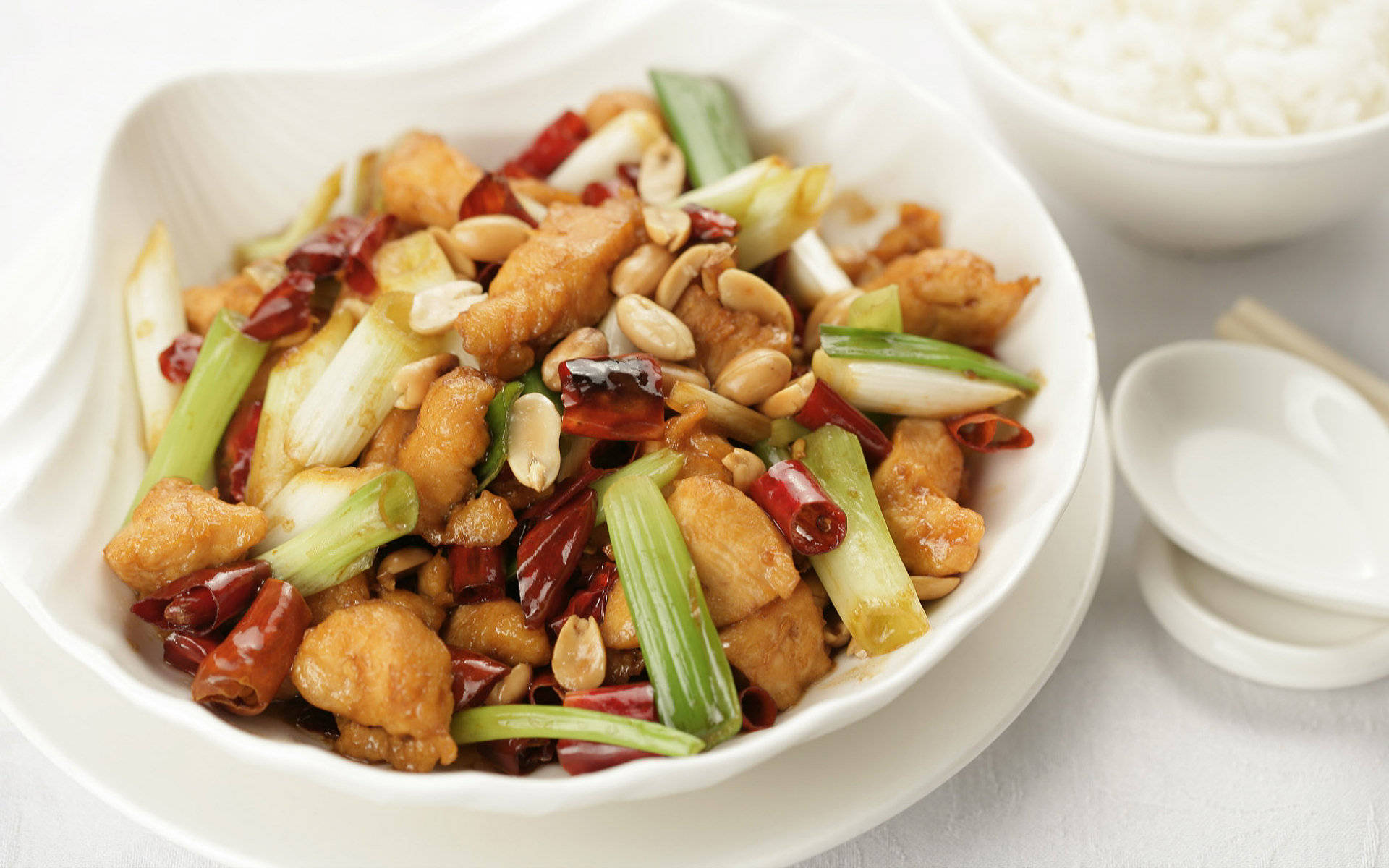 4K Food 桌布 壁紙 — 一碗色彩繽紛的宮保雞丁，配有花生、辣椒和青蔥，旁邊放著一碗白飯和筷子。