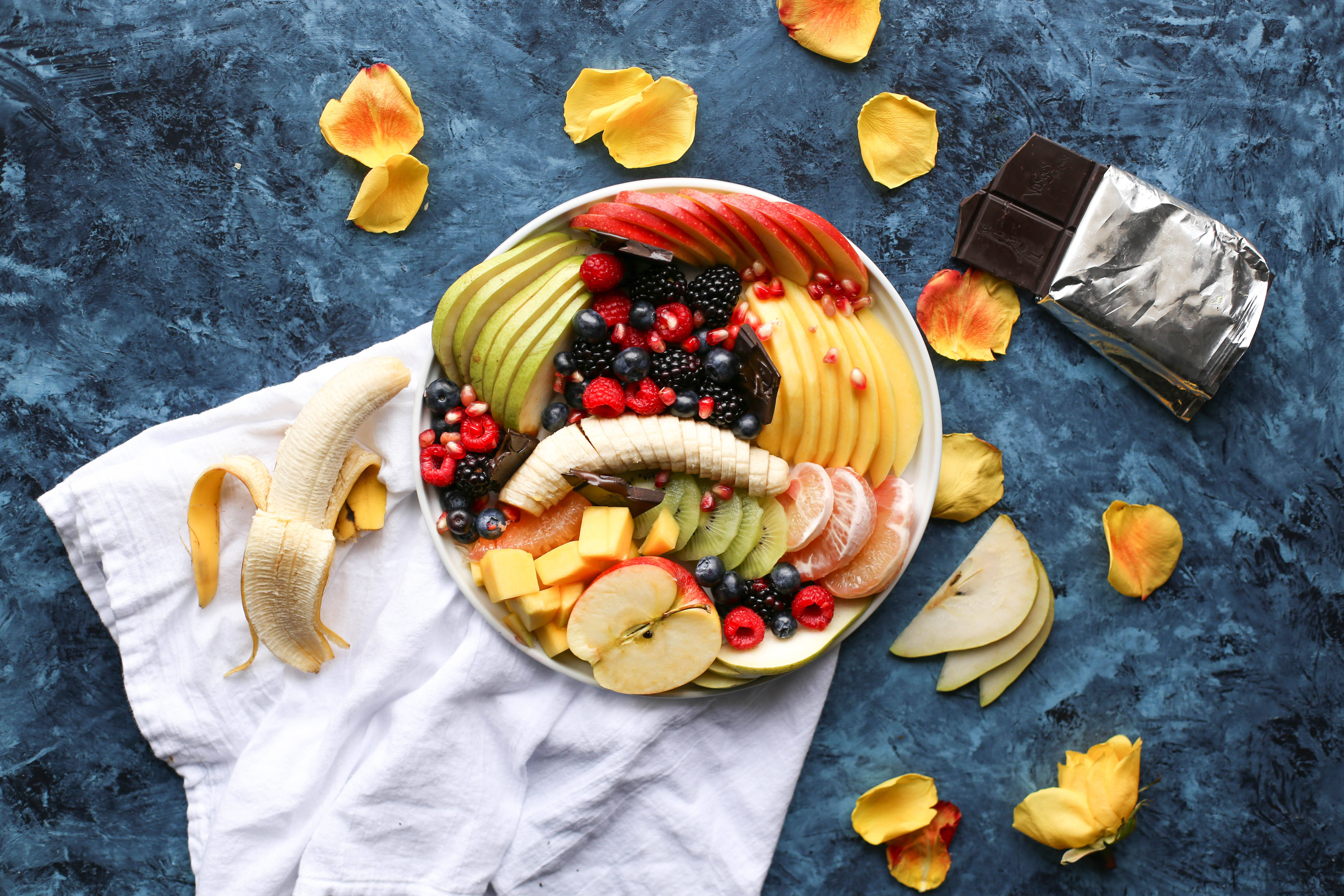 Fruit 桌布 壁紙 — 一碗色彩繽紛的新鮮水果，包括蘋果、香蕉和莓果，擺放在白色布上，背景為帶紋理的藍色，散落著黃色花瓣和一條巧克力。