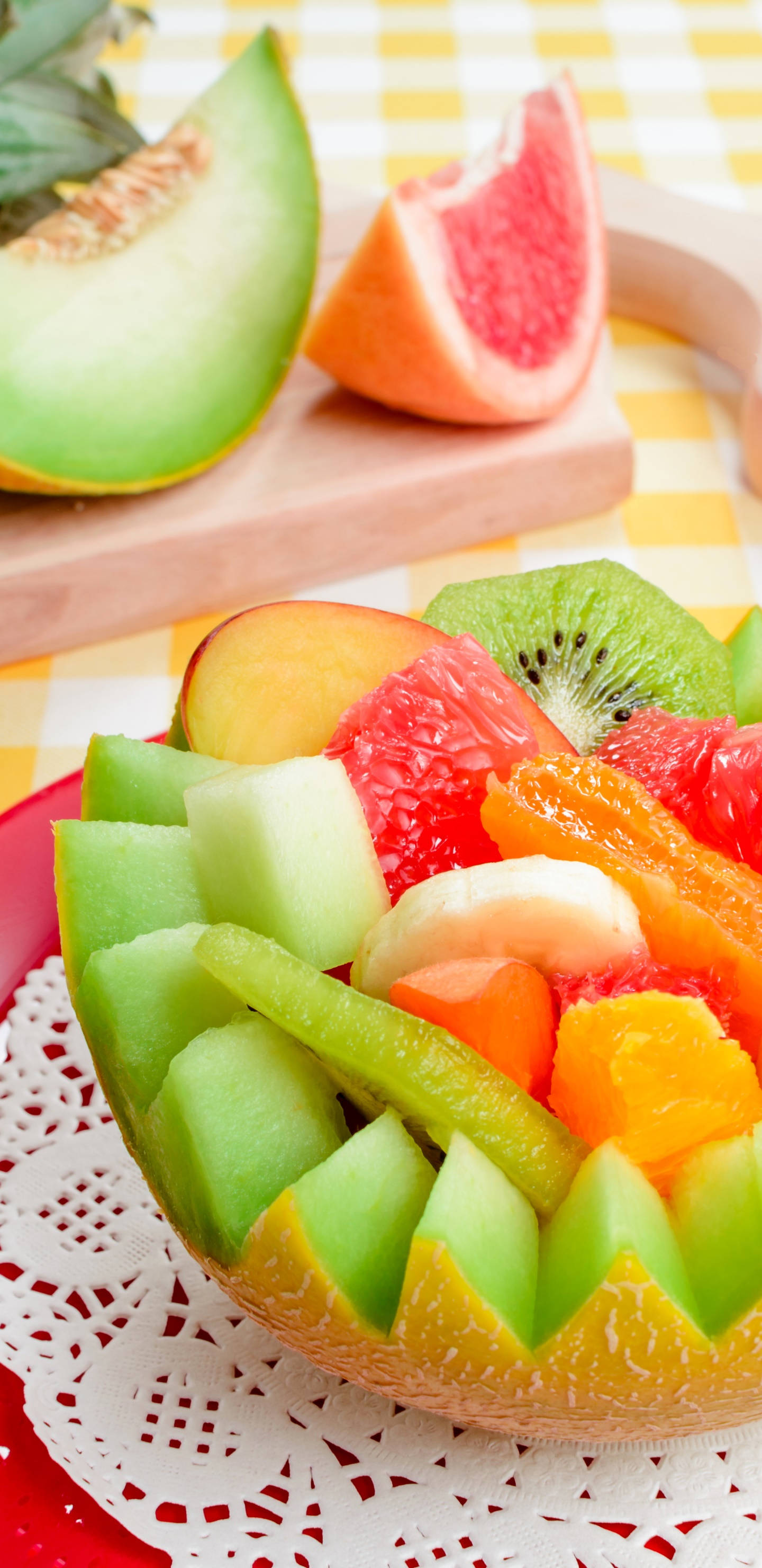 Fruits 桌布 壁紙 — 一碗色彩繽紛的新鮮水果，包括蜜瓜、奇異果、葡萄柚和柳橙，放在黃色格子桌布上。