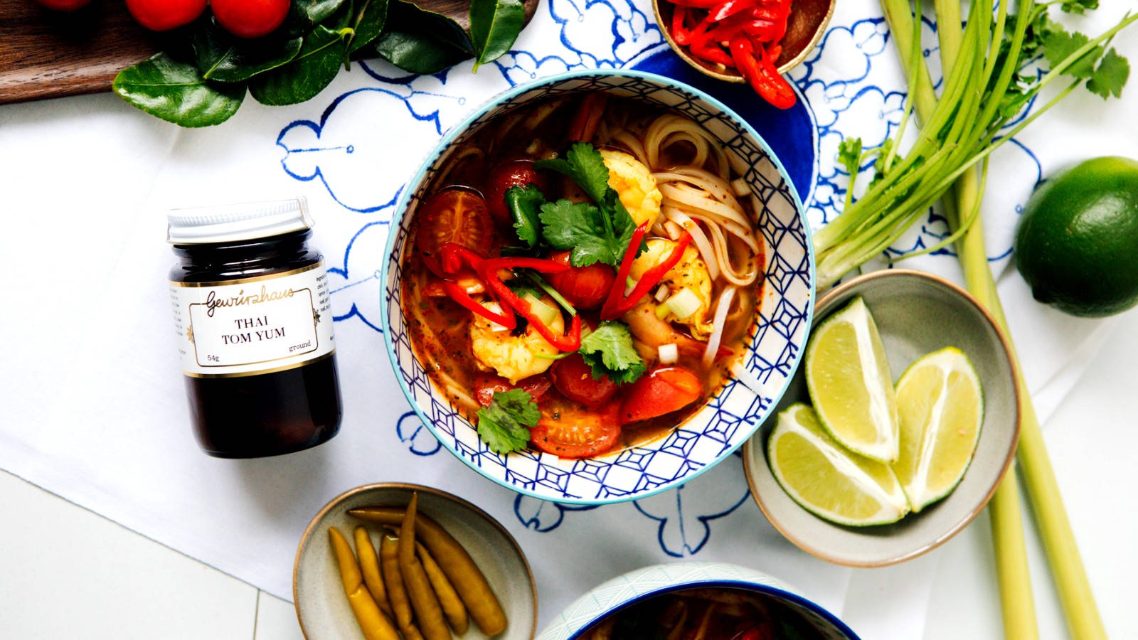 Food Photography 桌布 壁紙 — 一碗色彩繽紛的泰式冬陰功麵，配上蝦、辣椒和香菜，周圍環繞著新鮮青檸、香茅和其他食材。