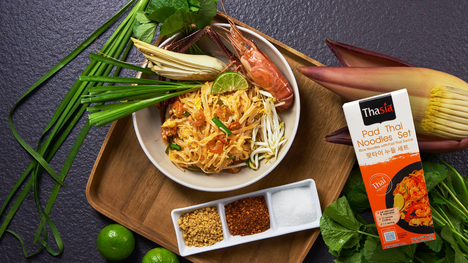 thai 桌布 壁紙 — 一碗色彩繽紛的泰式炒河粉，配上蝦、青檸和香草，放在木托盤上，旁邊是調味包和新鮮食材。