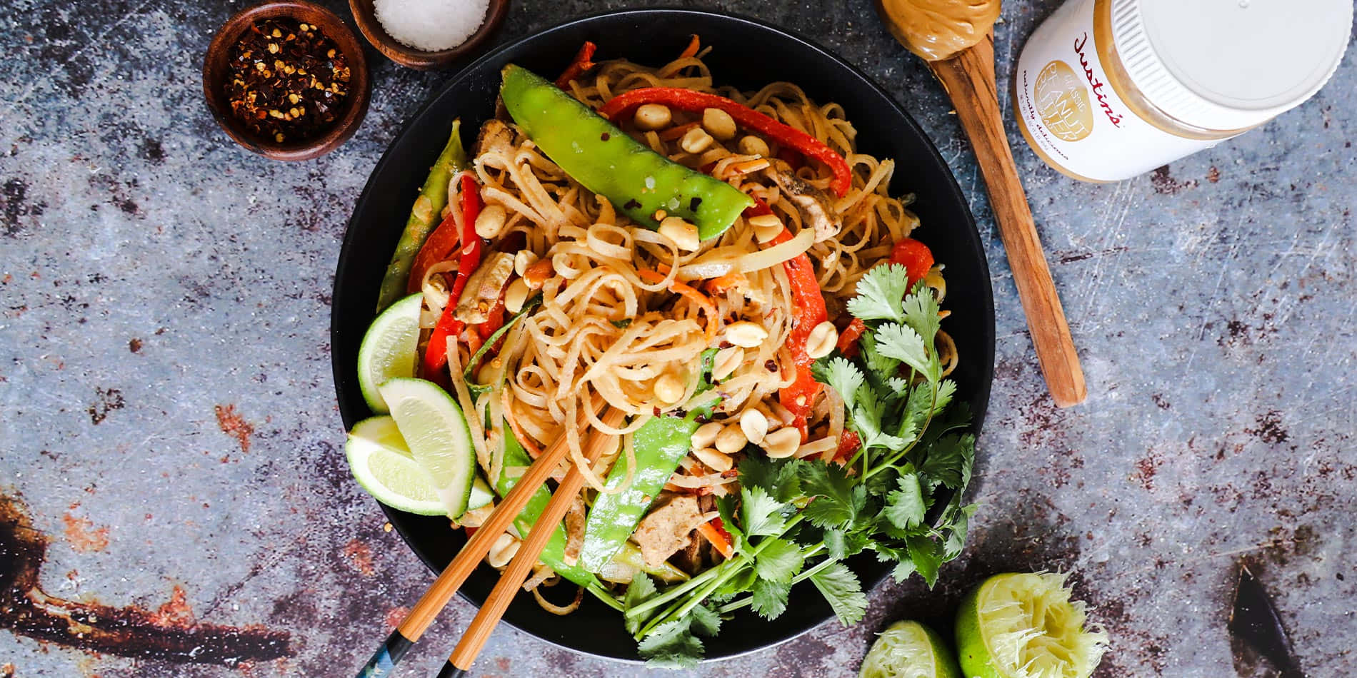 Noodles 桌布 壁紙 — 一碗色彩繽紛的炒麵，配上花生、辣椒、萊姆和新鮮香草，放在質樸的金屬表面上。
