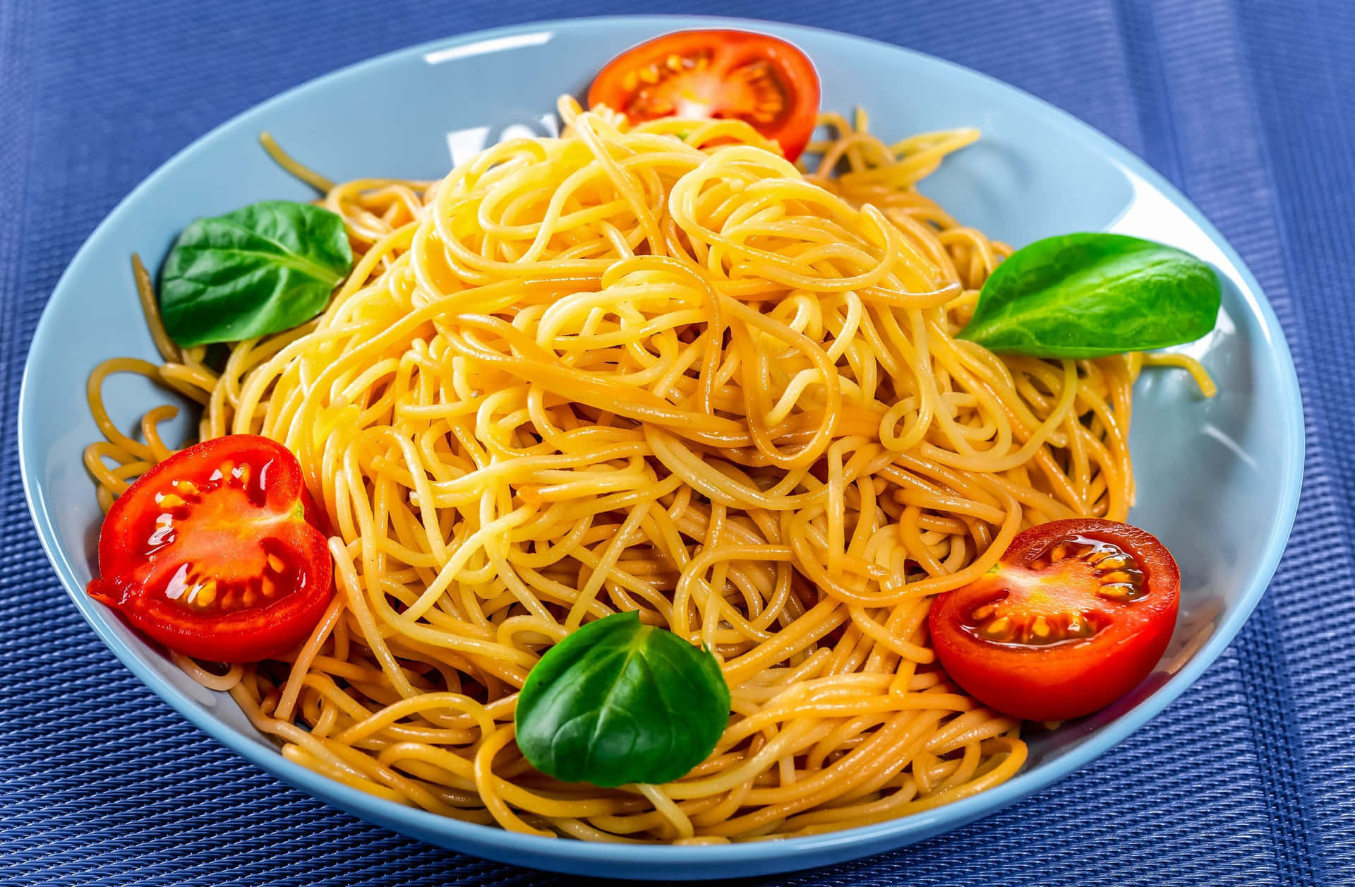 Noodles 桌布 壁紙 — 一碗色彩繽紛的義大利麵，上面放著新鮮羅勒和對半切開的櫻桃番茄，放在藍色紋理餐墊上。