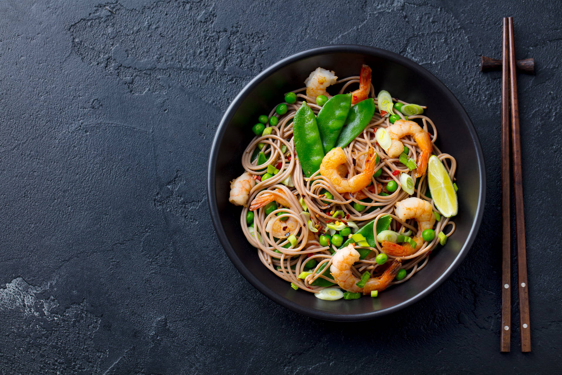 Noodles 桌布 壁紙 — 一碗色彩繽紛的蕎麥麵，配上蝦仁、荷蘭豆和青檸角，置於深色紋理表面上。