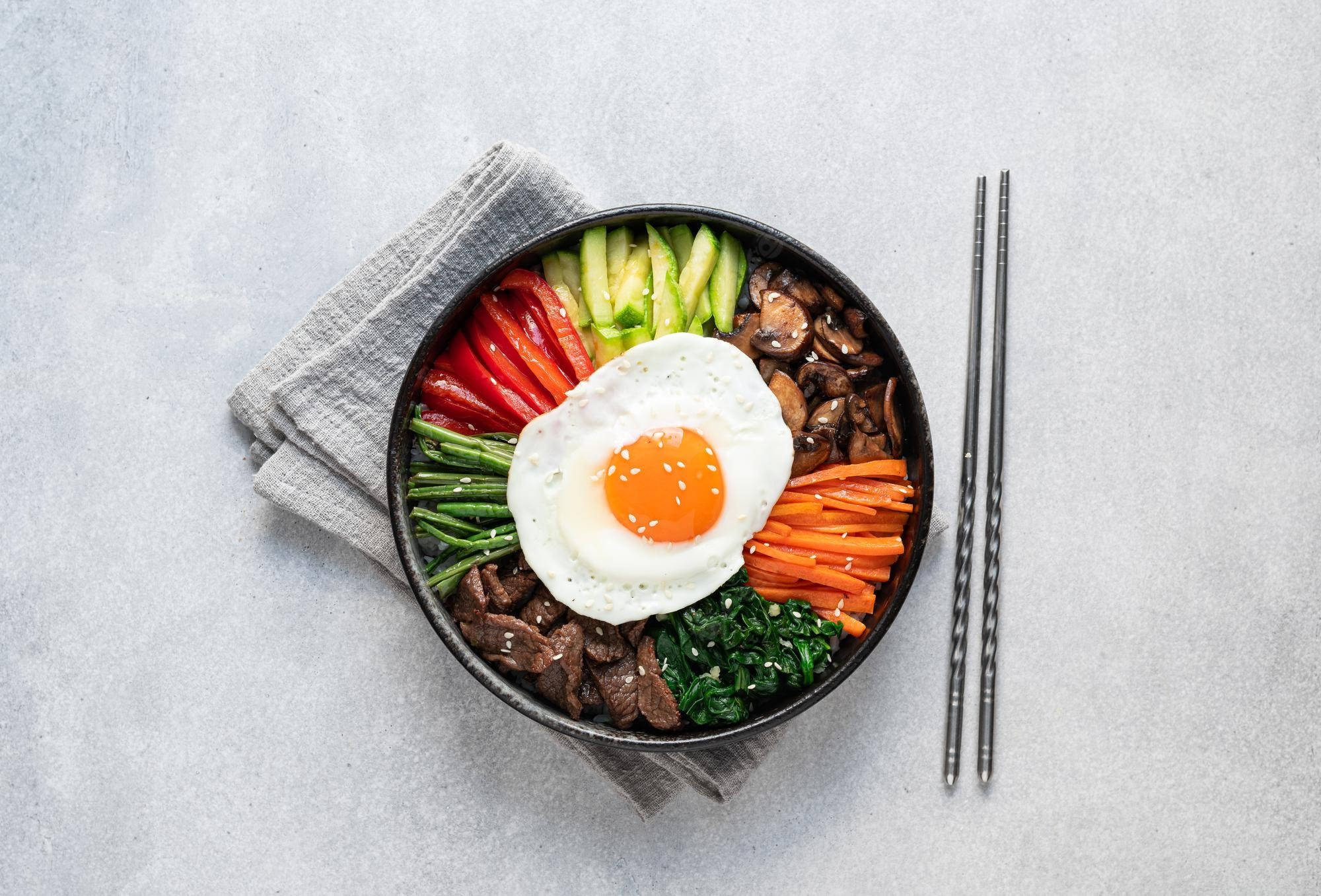烹飪 桌布 壁紙 — 一碗色彩繽紛的韓國石鍋拌飯，上面放著太陽蛋，放在灰色餐巾上，旁邊有筷子。