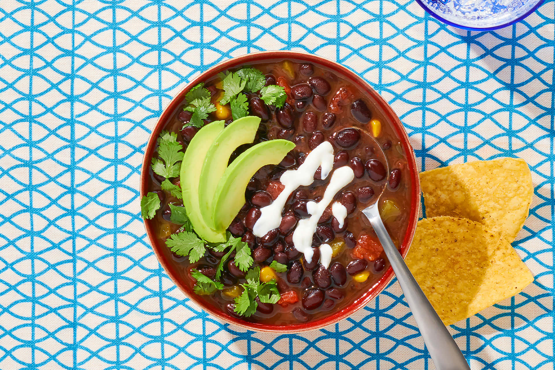 Black Bean Soup 桌布 壁紙 — 一碗色彩繽紛的黑豆辣椒，上面放著酪梨片、酸奶油和香菜，放在藍色圖案桌布上，附近有墨西哥玉米片。