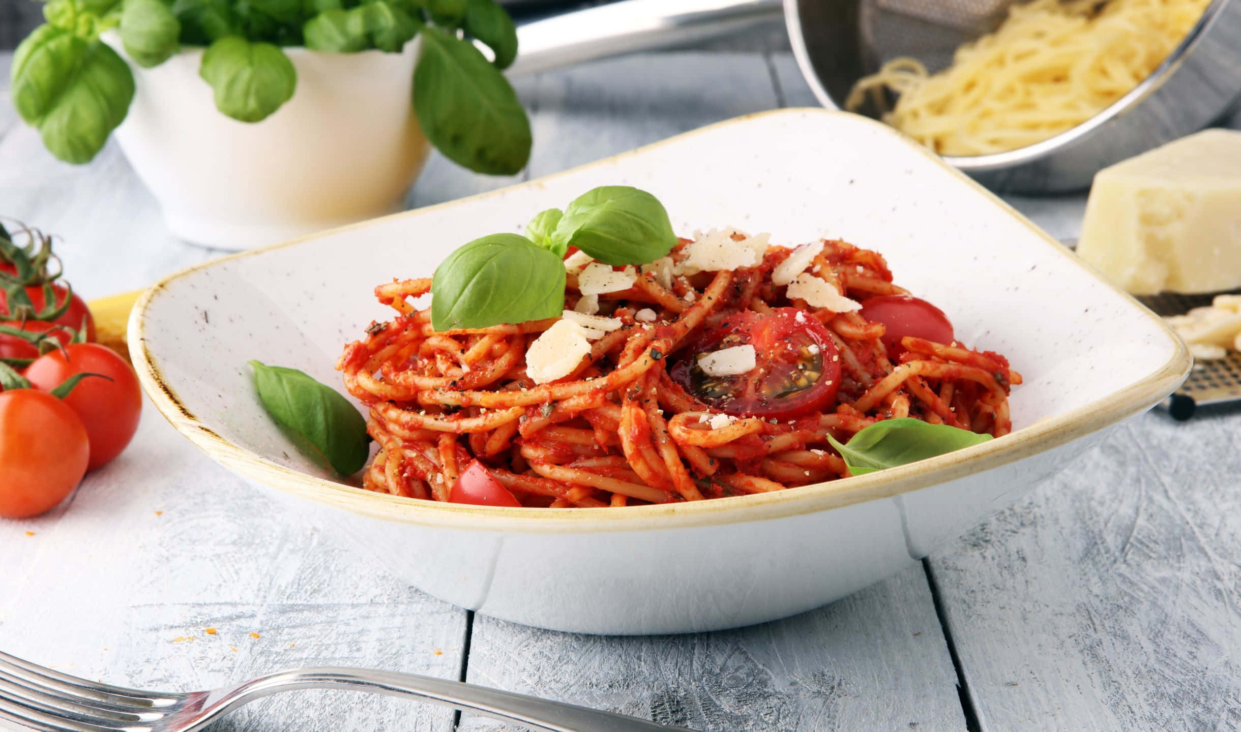 2440x1440 Pasta 桌布 壁紙 — 一碗色彩鮮豔的番茄義大利麵，配上羅勒和起司，放在鄉村風格的木桌上，旁邊還有新鮮食材。