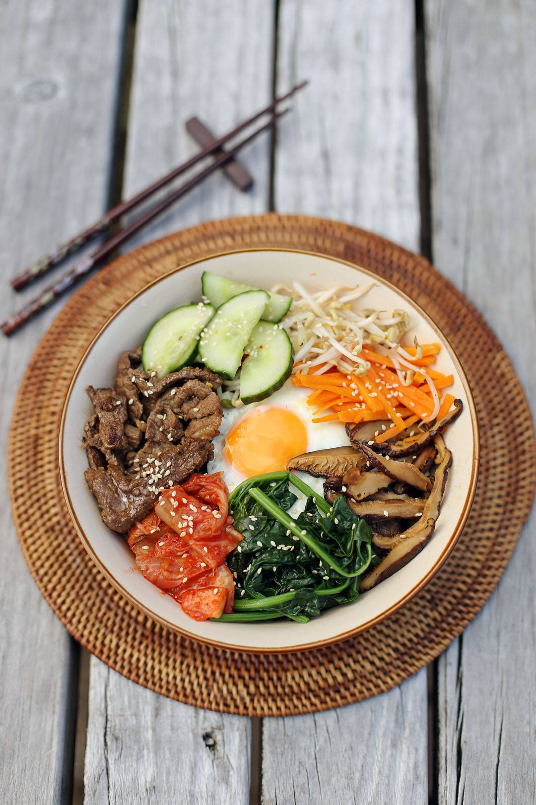 烹飪 桌布 壁紙 — 一碗色彩鮮豔的韓國石鍋拌飯，上面鋪有牛肉、雞蛋、蔬菜和芝麻，放在編織墊上，置於風化木頭上，旁邊有筷子。