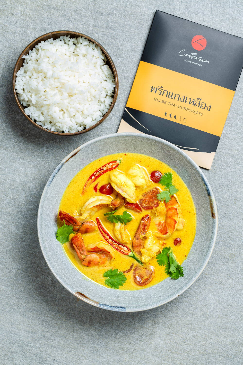 Delicious Food 桌布 壁紙 — 一碗色彩鮮豔的黃色泰式咖哩，搭配蝦和紅辣椒，旁邊放著一碗白飯和一個咖哩醬包，置於灰色表面上。