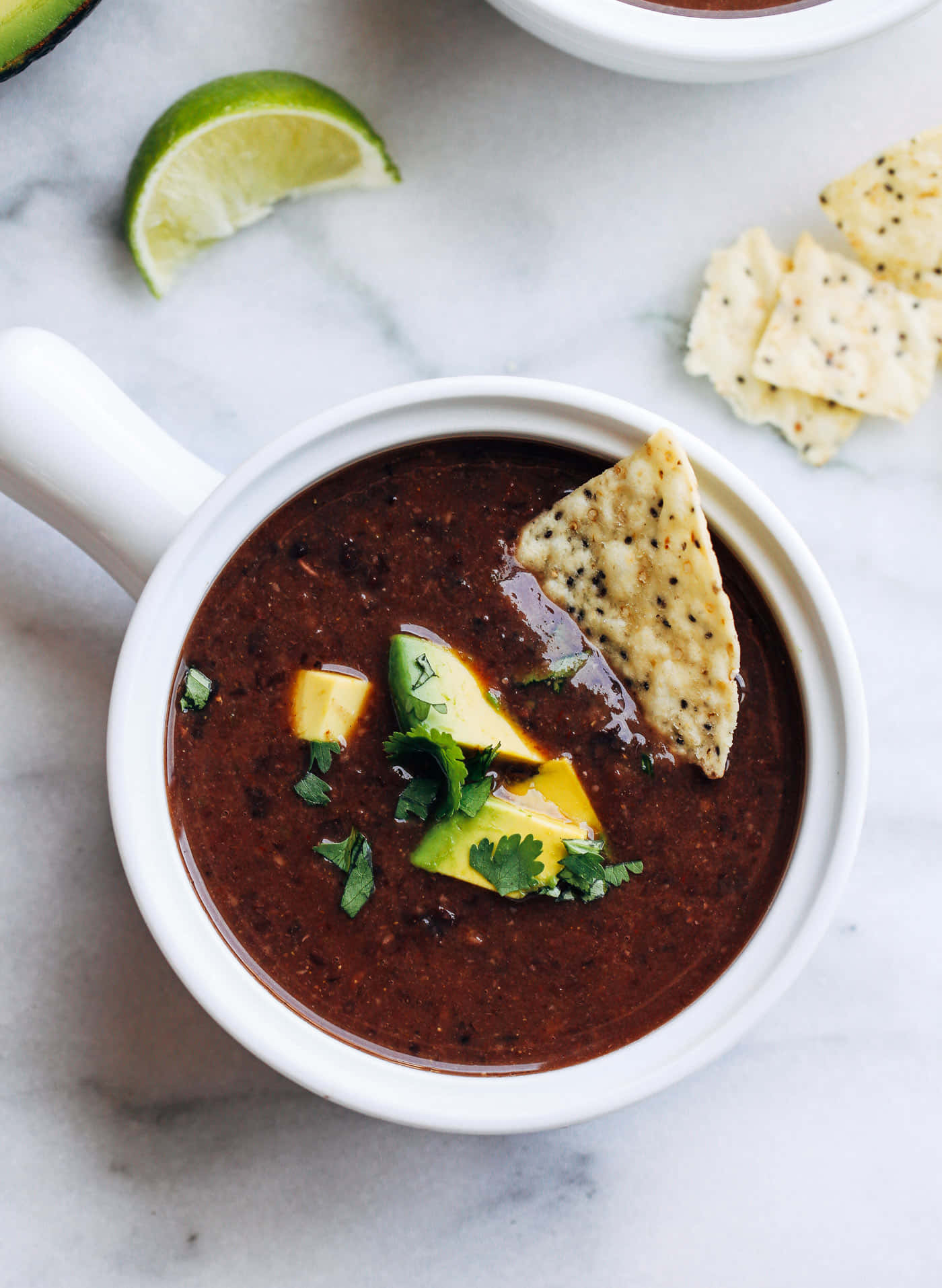 Black Bean Soup 桌布 壁紙 — 一碗豐富的黑豆湯，上面放有酪梨、萊姆和香菜，放在大理石表面上，旁邊有玉米片。