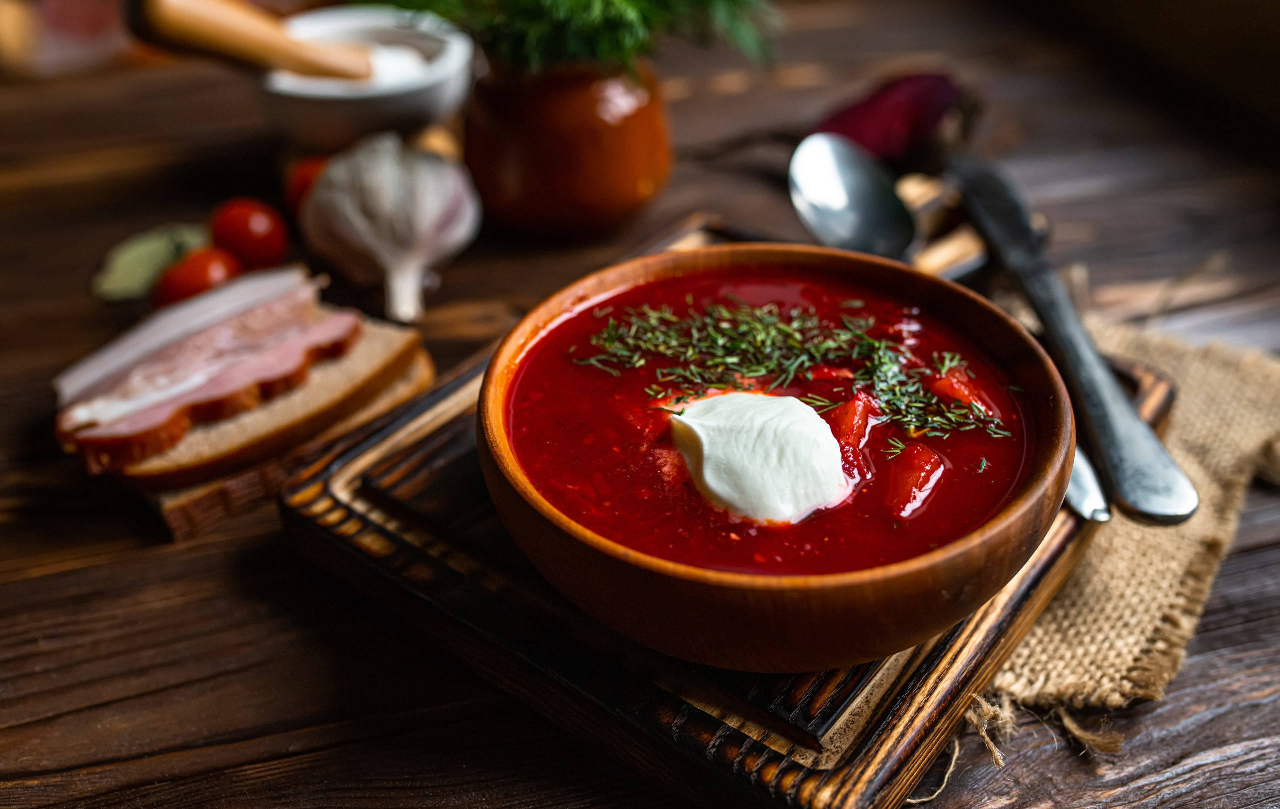 beetroot 桌布 壁紙 — 一碗豐盛的紅菜湯放在木碗裡，上面加了酸奶油和新鮮蒔蘿，旁邊有麵包和大蒜，擺在質樸的木桌上。