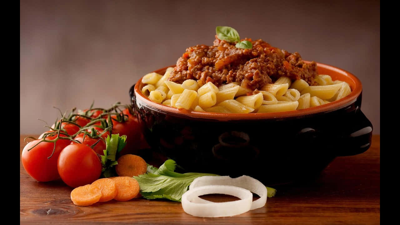 720p Pasta 桌布 壁紙 — 一碗豐盛的長通粉，配上濃郁的肉醬和新鮮羅勒，旁邊還有鮮豔的櫻桃番茄、紅蘿蔔和洋蔥，放在質樸的木桌上。