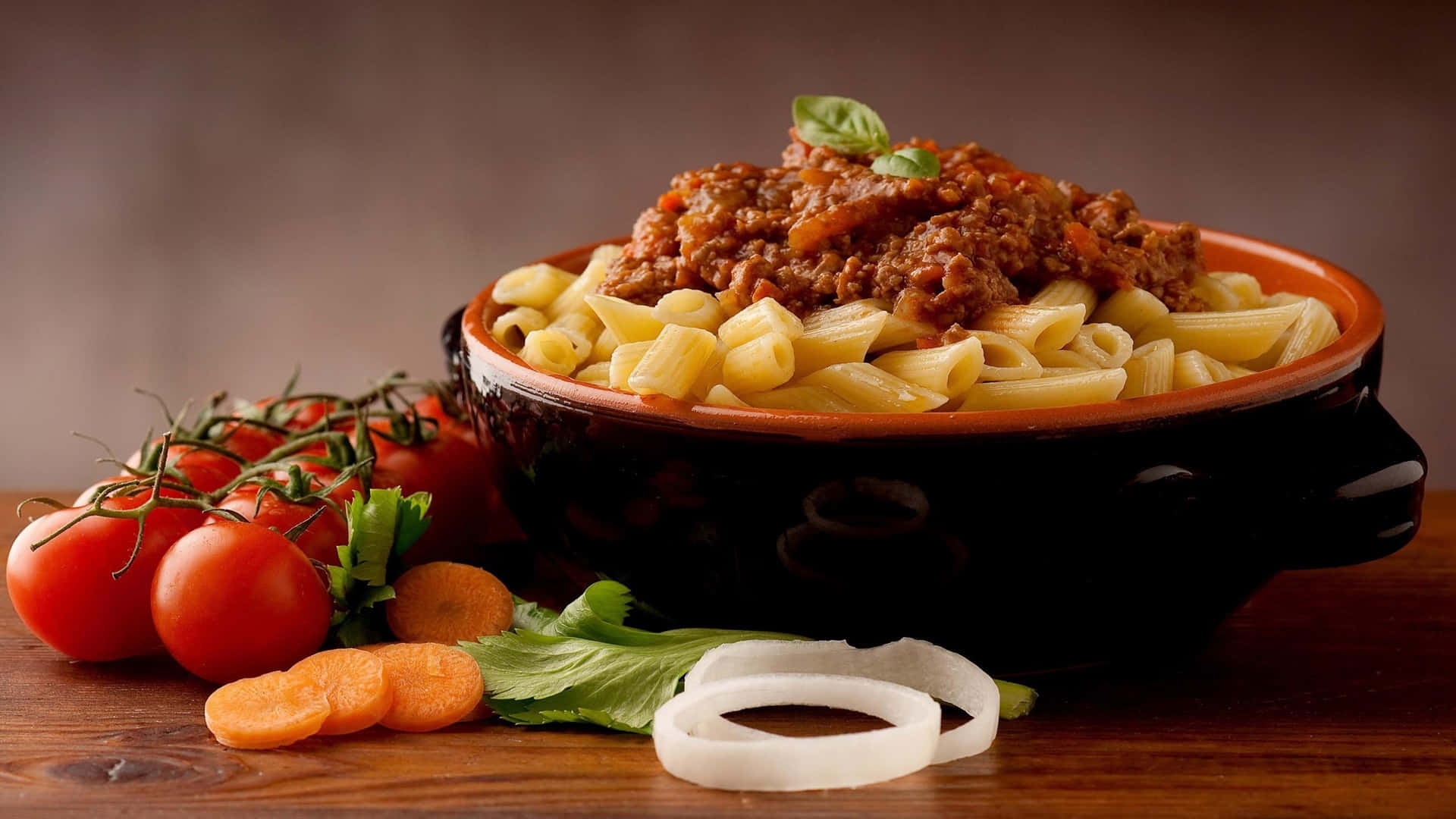 Italian 桌布 壁紙 — 一碗鄉村風格的筆管麵，淋上濃郁肉醬，並以新鮮羅勒點綴，旁邊放著鮮豔的小番茄、紅蘿蔔和洋蔥，置於木桌上。
