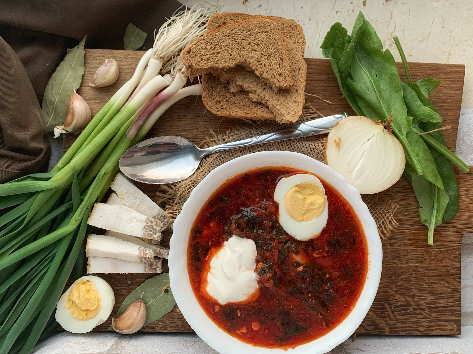 beetroot 桌布 壁紙 — 一碗鄉村風格的鮮紅色羅宋湯，上面放有酸奶油和水煮蛋，放在木板上，周圍有蒜頭、青蔥和黑麥麵包等新鮮食材。