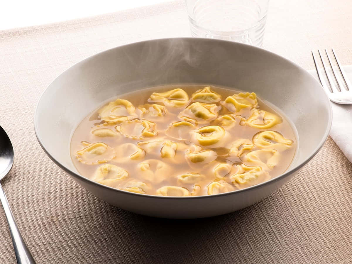 Food Photography 桌布 壁紙 — 一碗金黃色義大利餃子在奶油湯中冒著蒸氣，放在有紋理的米色桌布上，旁邊有餐具。