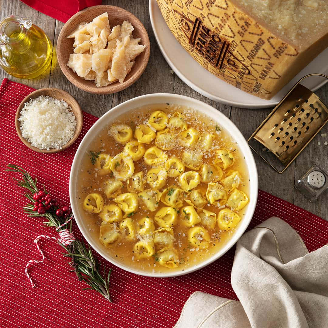 Food Photography 桌布 壁紙 — 一碗金黃色義大利餃子在湯中，配上香草和起司，放在紅色布上，旁邊有帕馬森起司、刨絲器和橄欖油。
