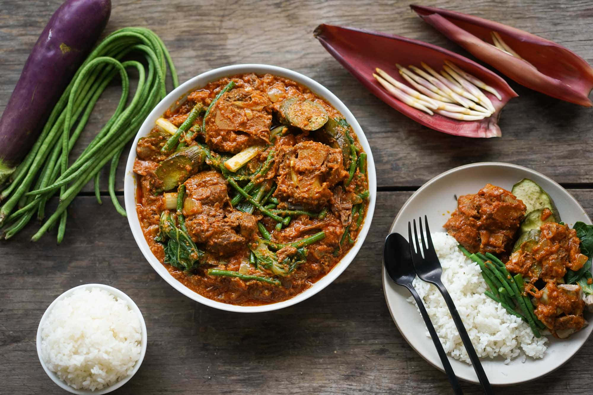 meat 桌布 壁紙 — 一碗香料豐富的咖哩，配有肉類和青豆，盛載於白色碗中，旁邊有米飯和新鮮食材，置於鄉村風格的木桌上。