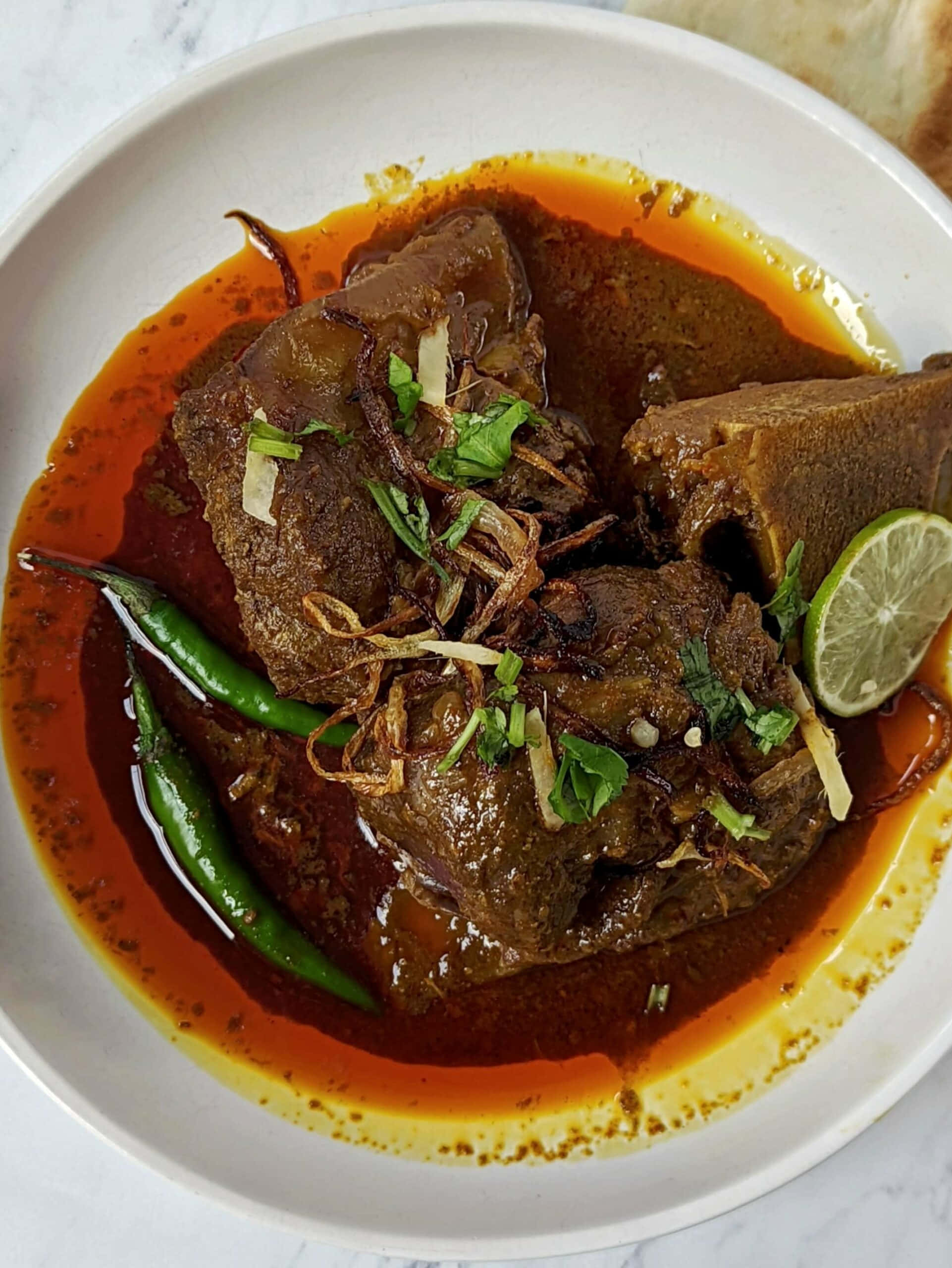 Nihari 桌布 壁紙 — 一碗香料豐富的羊肉咖哩，配上青辣椒、萊姆和炸洋蔥，在白色碗中閃閃發光。
