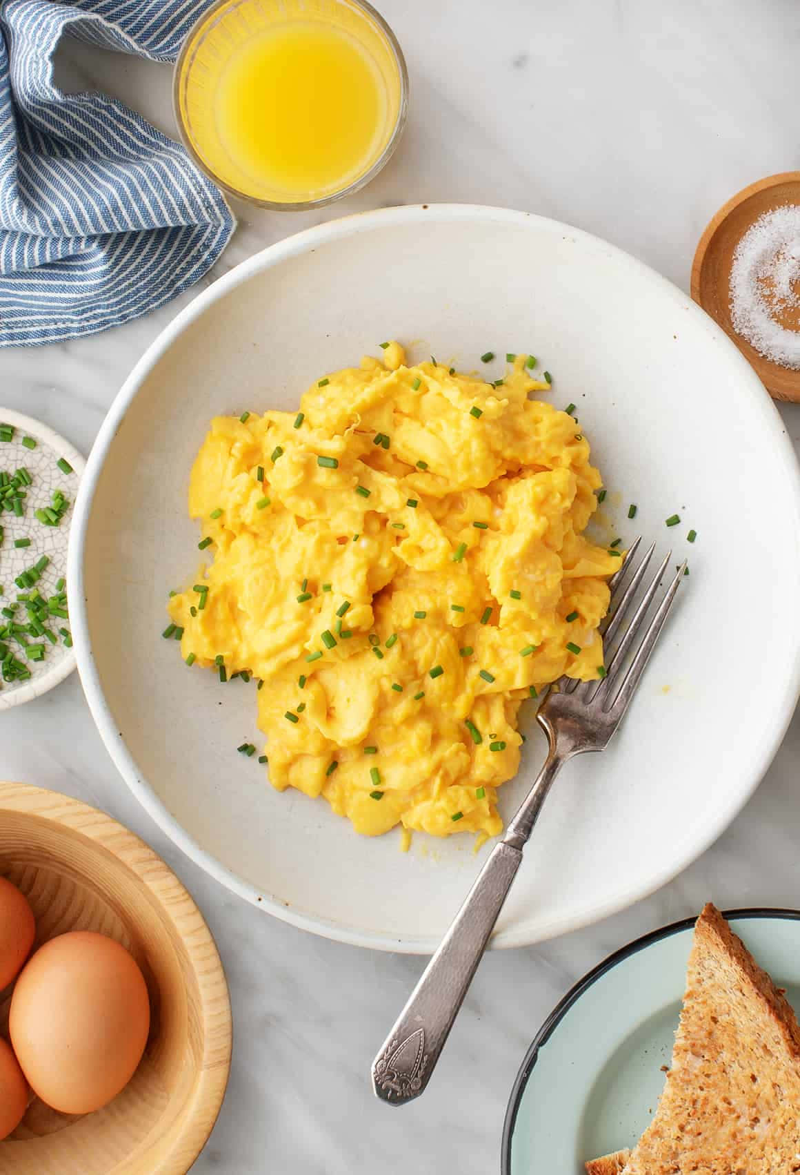 Scrambled Egg 桌布 壁紙 — 一碗鬆軟金黃色的炒蛋，上面撒上切碎的細香蔥，放在大理石檯面上，旁邊有吐司和柳橙汁。