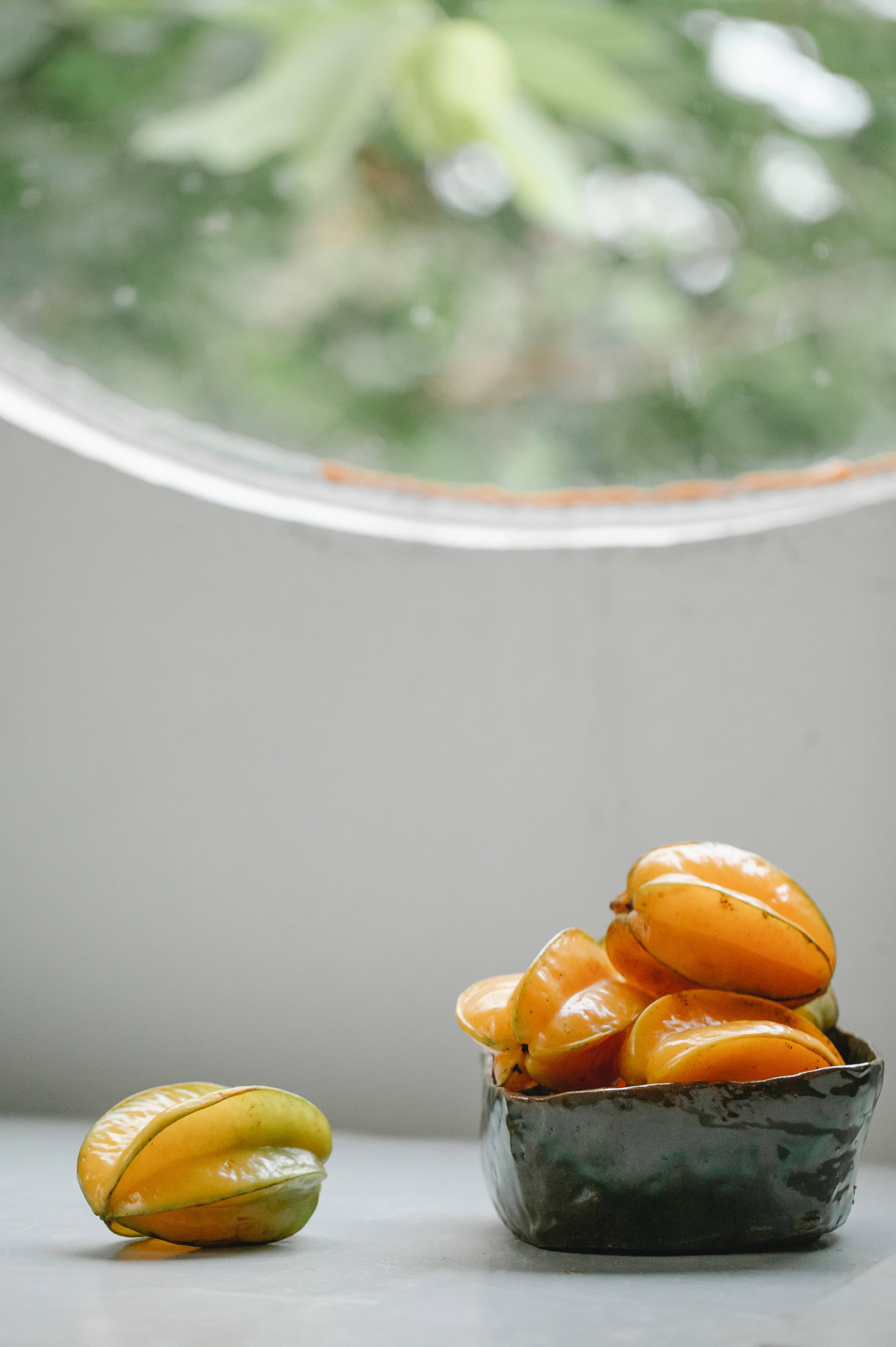 Fruit 桌布 壁紙 — 一碗鮮橙色楊桃放在灰色表面上，旁邊還有一個水果，柔和自然光從圓窗灑落。