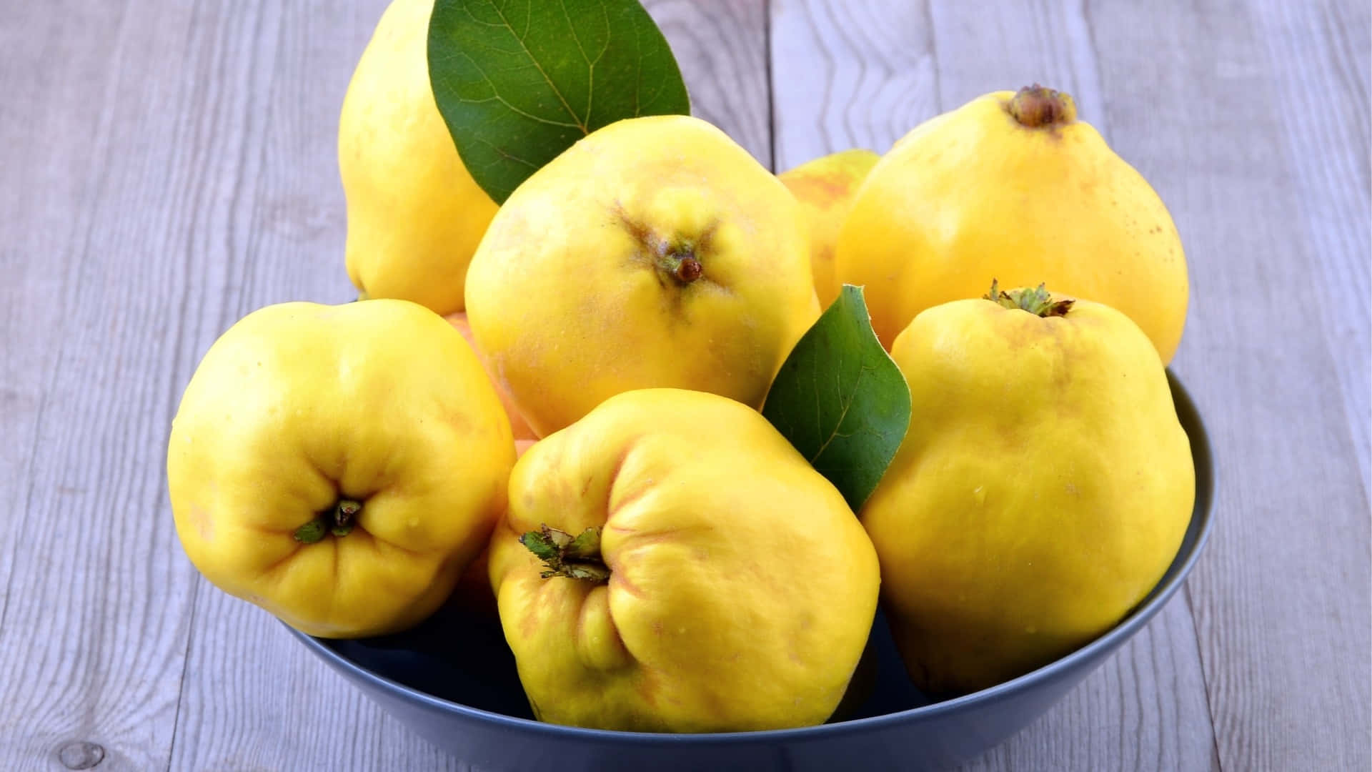 Quince 桌布 壁紙 — 一碗鮮黃色的榲桲配綠葉，放在經風化的木質表面上。