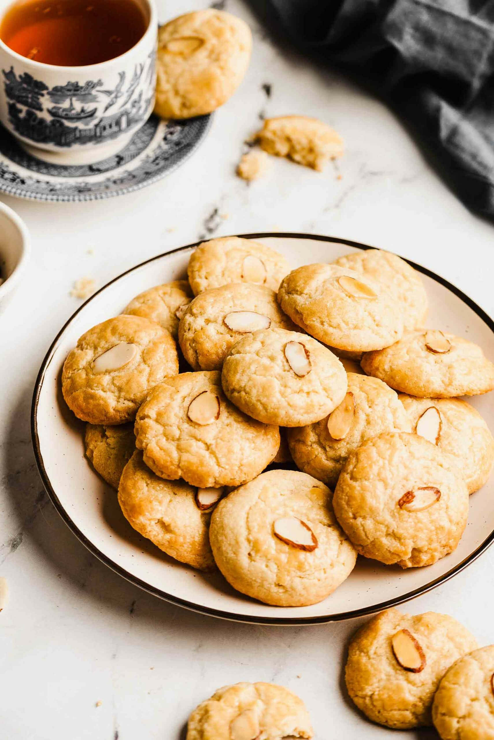 Cookie 桌布 壁紙 — 一碟金黃色的杏仁餅乾放在一杯茶旁邊，置於大理石表面上，附近有一塊深色布料。