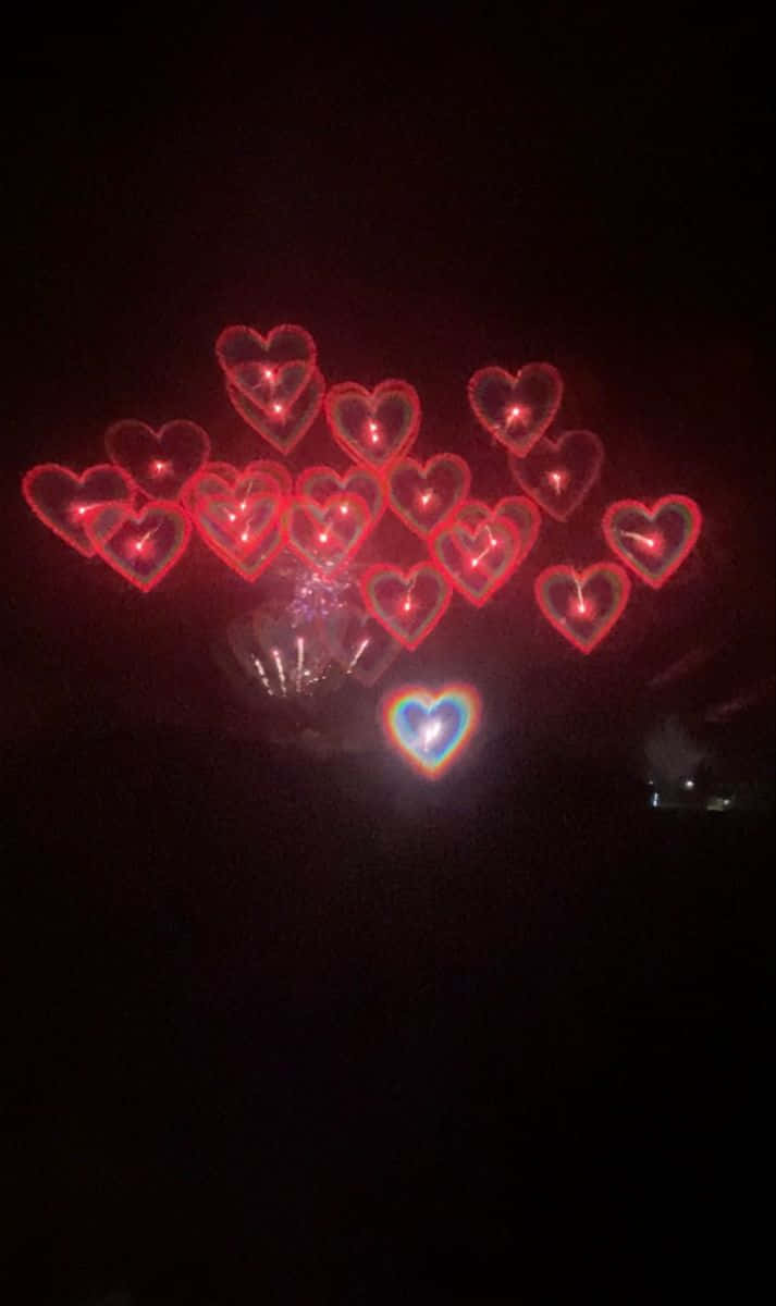 心形美學 桌布 壁紙 — 一簇發光的紅色心形煙火在黑暗的夜空中綻放，下方有一顆彩虹色的心。