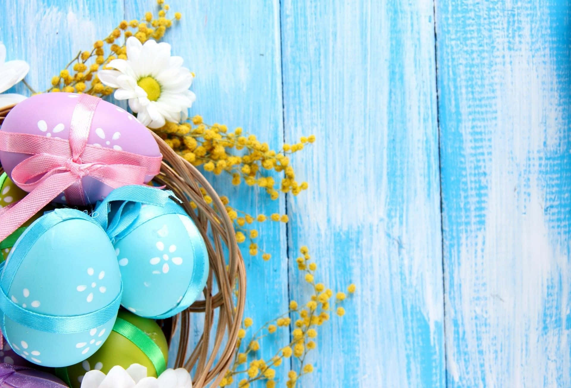 Pastel Easter 桌布 壁紙 — 一籃色彩繽紛的復活節彩蛋，飾有絲帶和雛菊，放在經風化的藍色木質表面上。