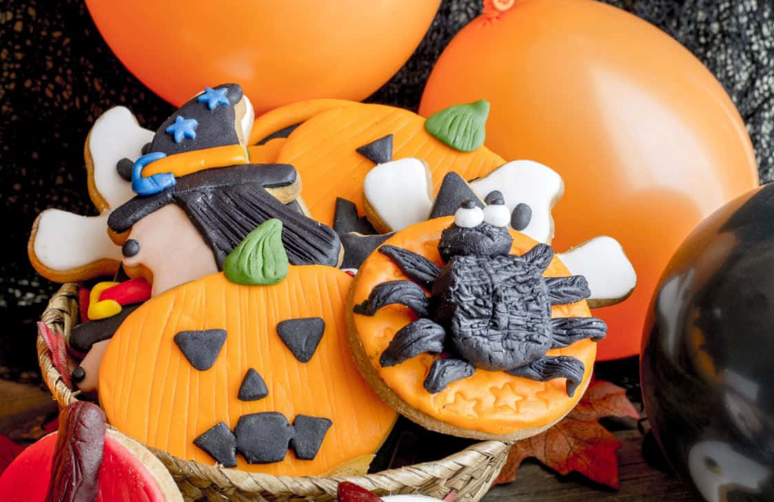 Halloween Treats 桌布 壁紙 — 一籃萬聖節主題糖餅乾，形狀有南瓜、女巫和蜘蛛，周圍環繞著橙色氣球和秋葉。