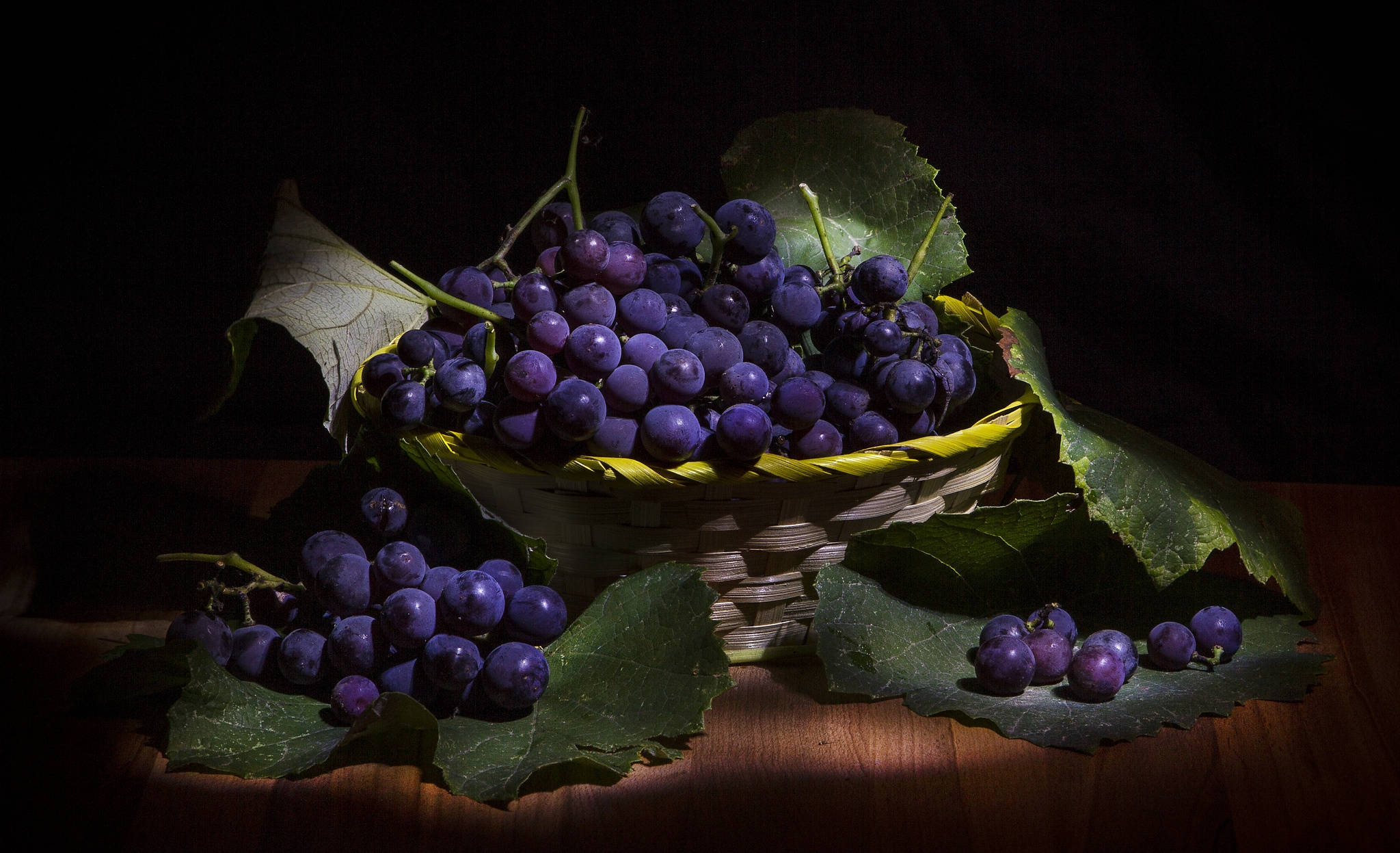 Grape 桌布 壁紙 — 一籃飽滿的紫葡萄配綠葉，放在木質表面上，在黑色背景下的戲劇性聚光燈照射下。
