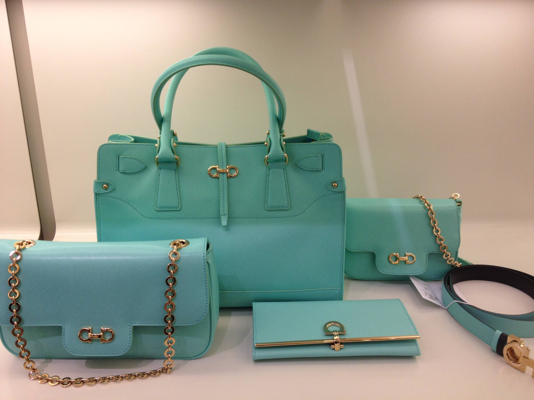 Ferragamo 桌布 壁紙 — 一系列優雅的綠松石色手提包及配件，搭配金色五金配件，展示在潔淨的白色表面上。