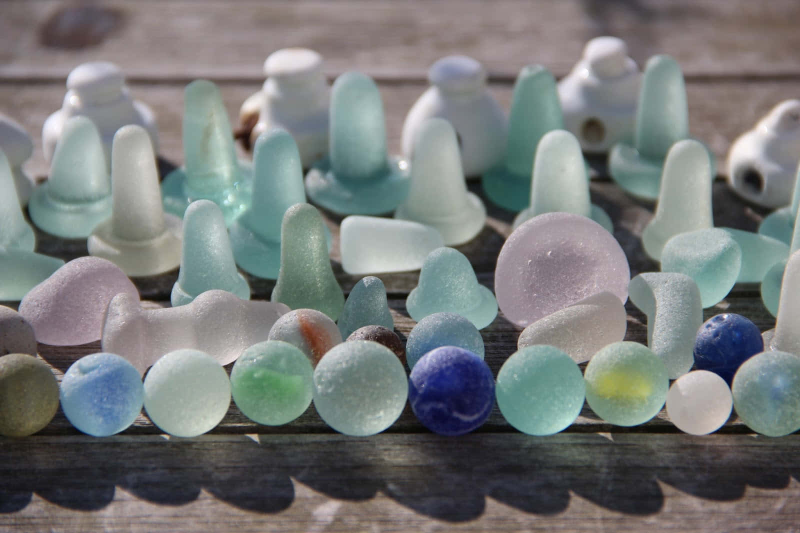 Sea Glass 桌布 壁紙 — 一系列光滑的海玻璃瓶和球體，以粉藍、粉紅和白色等柔和色調，排列在經風化的木板上，沐浴在明亮的陽光下。