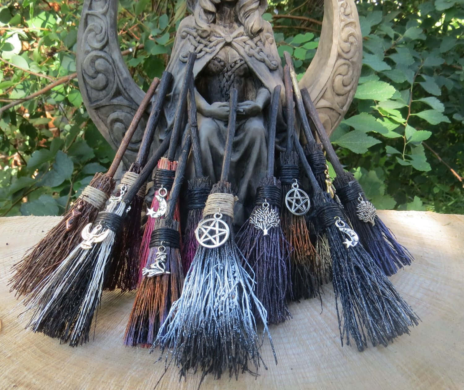 Witches Broom 桌布 壁紙 — 一系列帶有金屬飾物的華麗女巫掃帚，放在雕刻女巫雕像前，周圍是茂密的綠色植物。