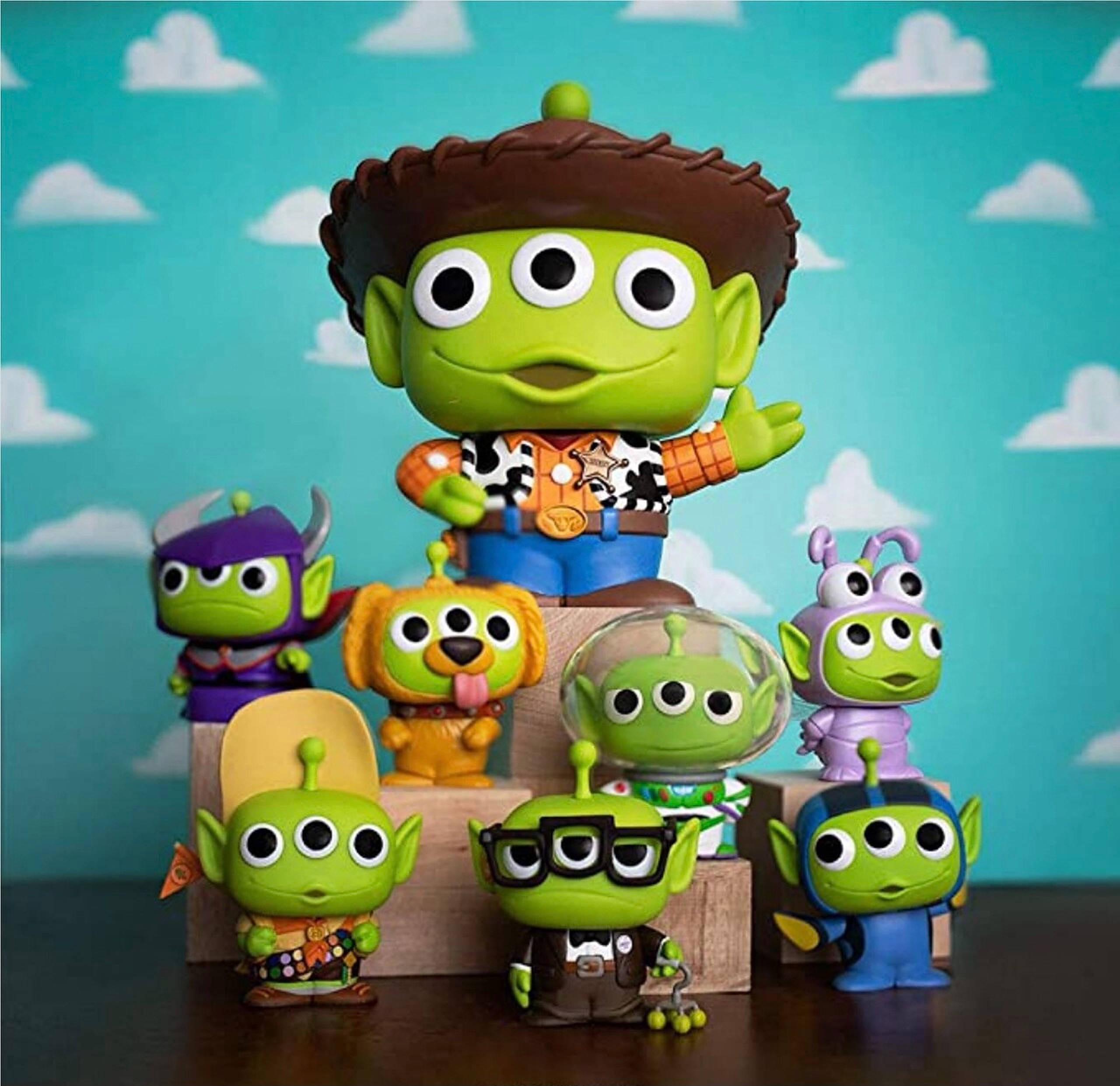 Toy Story Alien 桌布 壁紙 — 一系列綠色三眼外星人公仔，穿著各種服裝，擺放在木塊上，背景是帶有白雲的藍綠色天空。