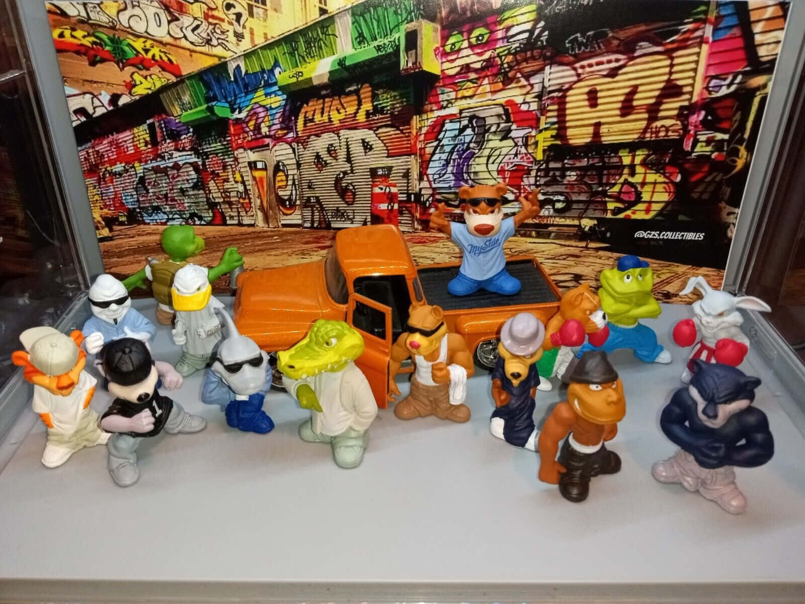 toys 桌布 壁紙 — 一系列色彩繽紛的卡通動物公仔，圍繞著一輛橙色卡車，背景是充滿活力的塗鴉牆。