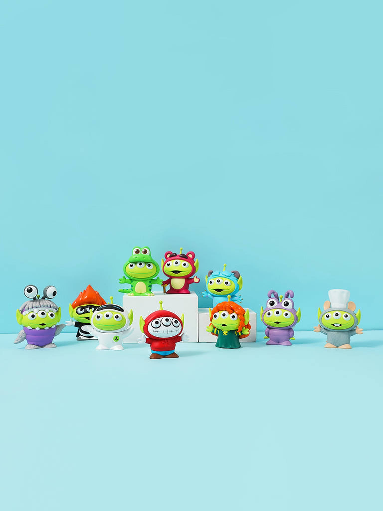 Toy Story Alien 桌布 壁紙 — 一系列色彩繽紛的小型綠色外星人玩具公仔，穿著各種服裝，排列在白色積木上，背景為純淺藍色。