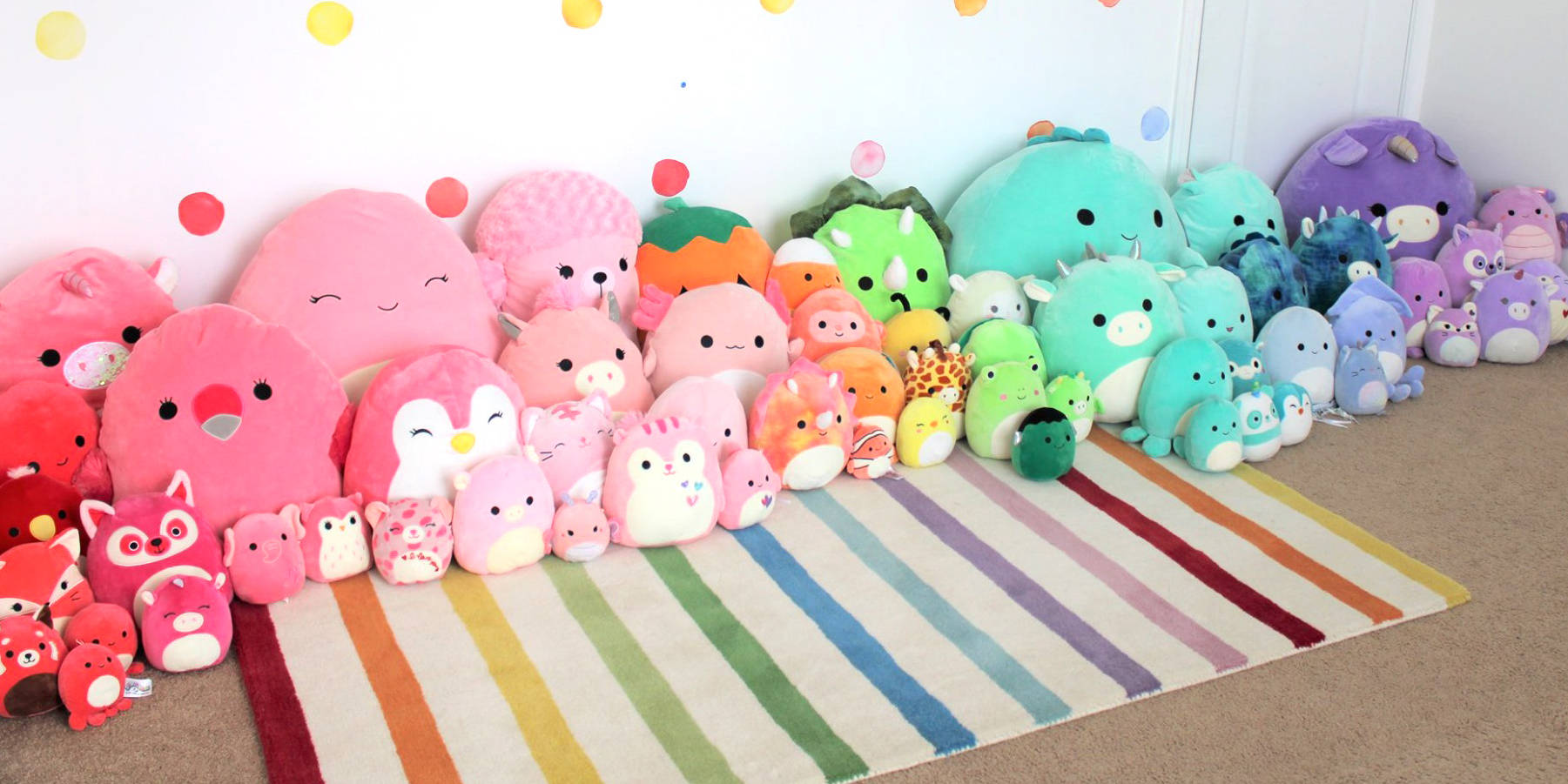 一張彩虹地毯，上面有一排色彩繽紛的毛絨玩具，背景為圓點牆。 — Squishmallows 壁紙
