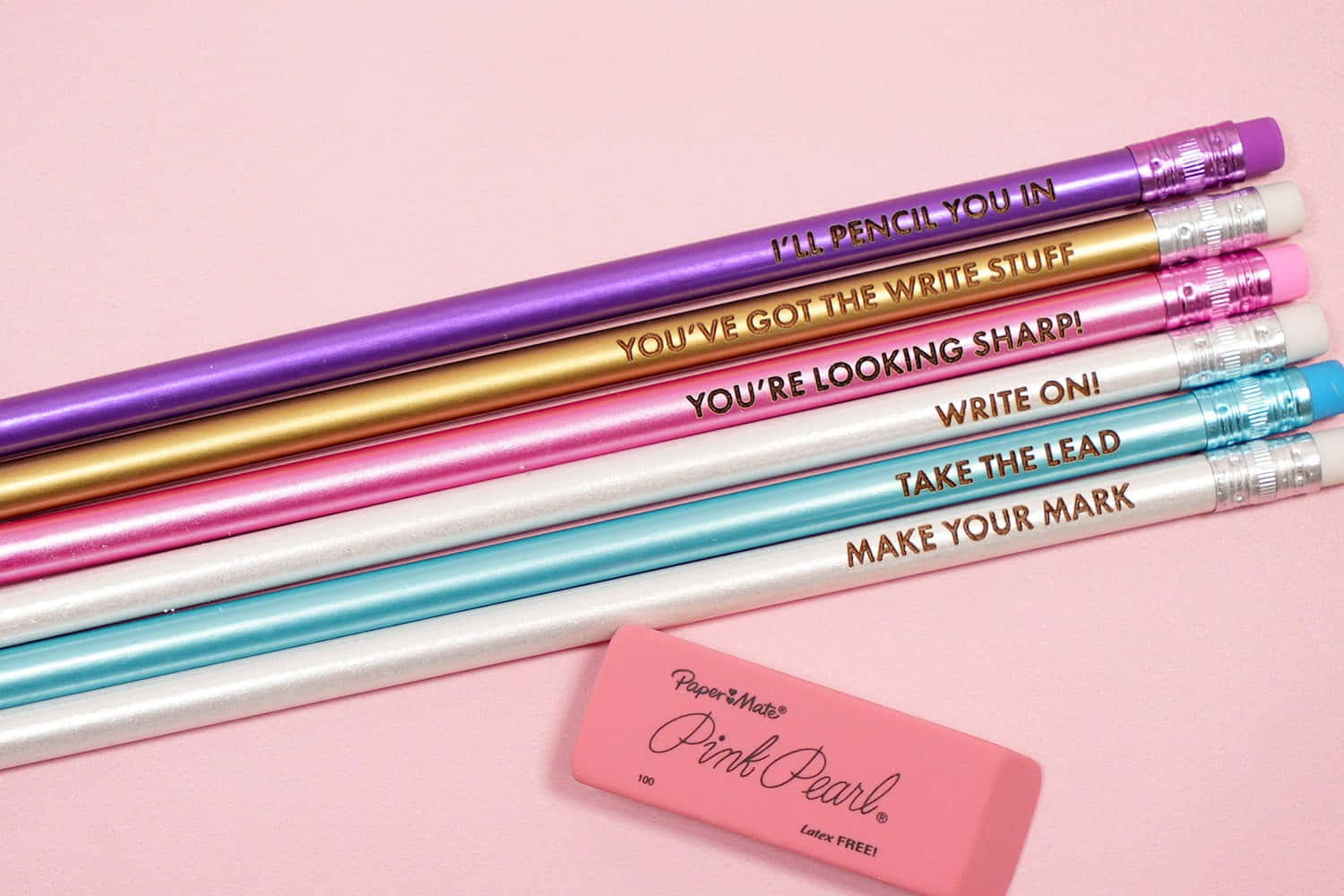 pencil 桌布 壁紙 — 一系列色彩繽紛的金屬鉛筆，上面印有勵志語句，斜放在粉紅色軟墊背景上的粉紅色橡皮擦旁邊。