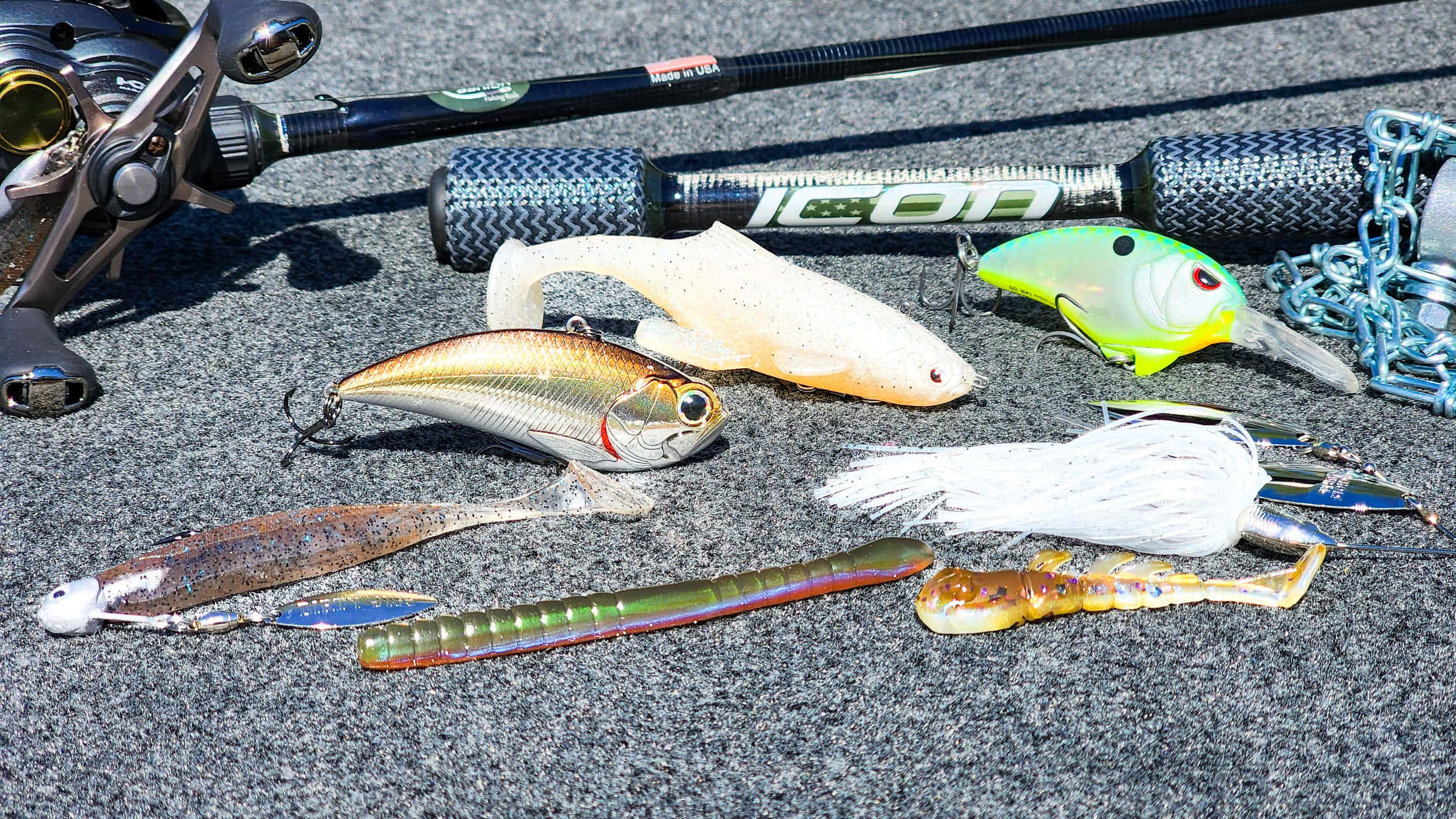 Fishing Bait 桌布 壁紙 — 一系列色彩繽紛的魚餌和一支釣竿放在有紋理的灰色瀝青上，在明亮的陽光下。