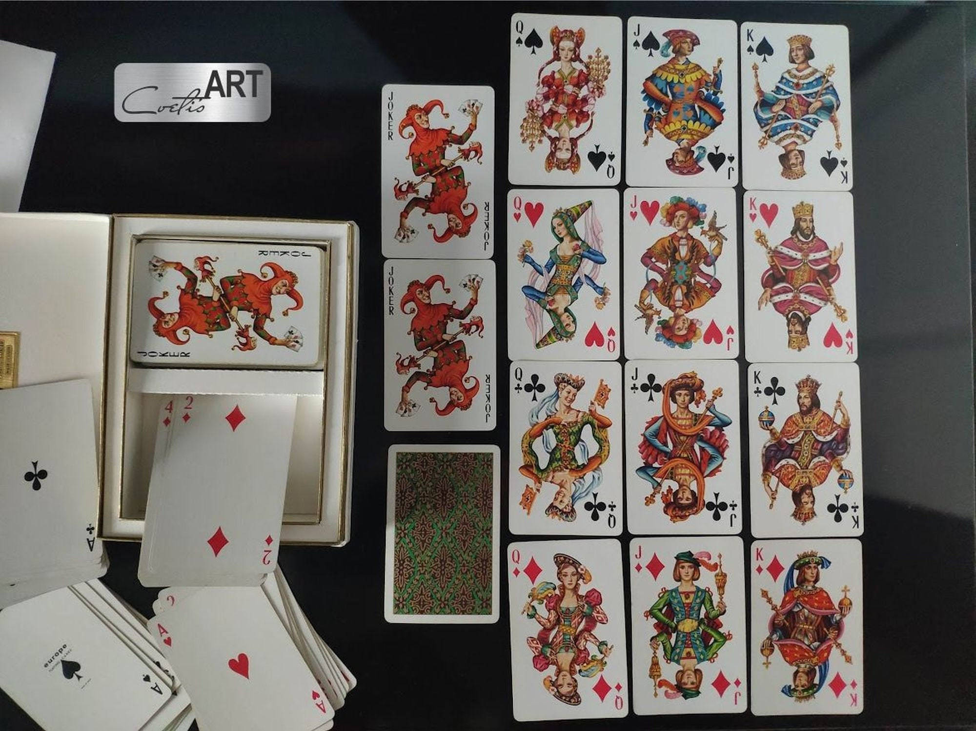 Playing Cards 桌布 壁紙 — 一系列裝飾華麗的撲克牌，上面有色彩鮮明的皇室人物和小丑，排列在深色表面上，旁邊有一個打開的牌盒。