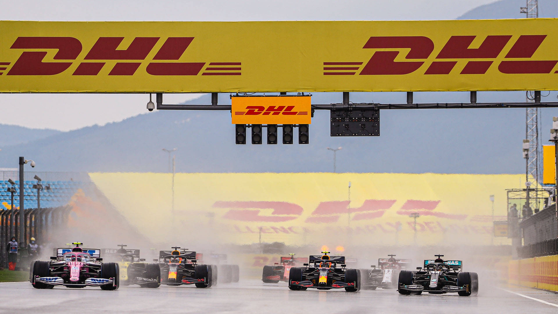 Dhl 桌布 壁紙 — 一級方程式賽車在濕滑賽道上緊密競賽，大型黃色DHL橫幅下輪胎升起霧氣。