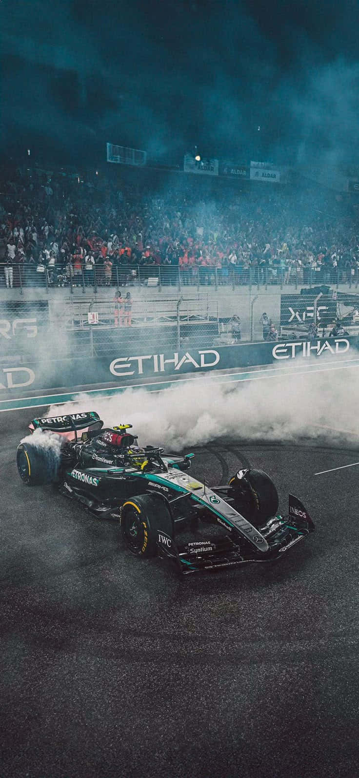 Mercedes F1 壁紙 — 一級方程式賽車在賽道上戲劇性地冒煙，人群在深藍色天空下的看台上歡呼。